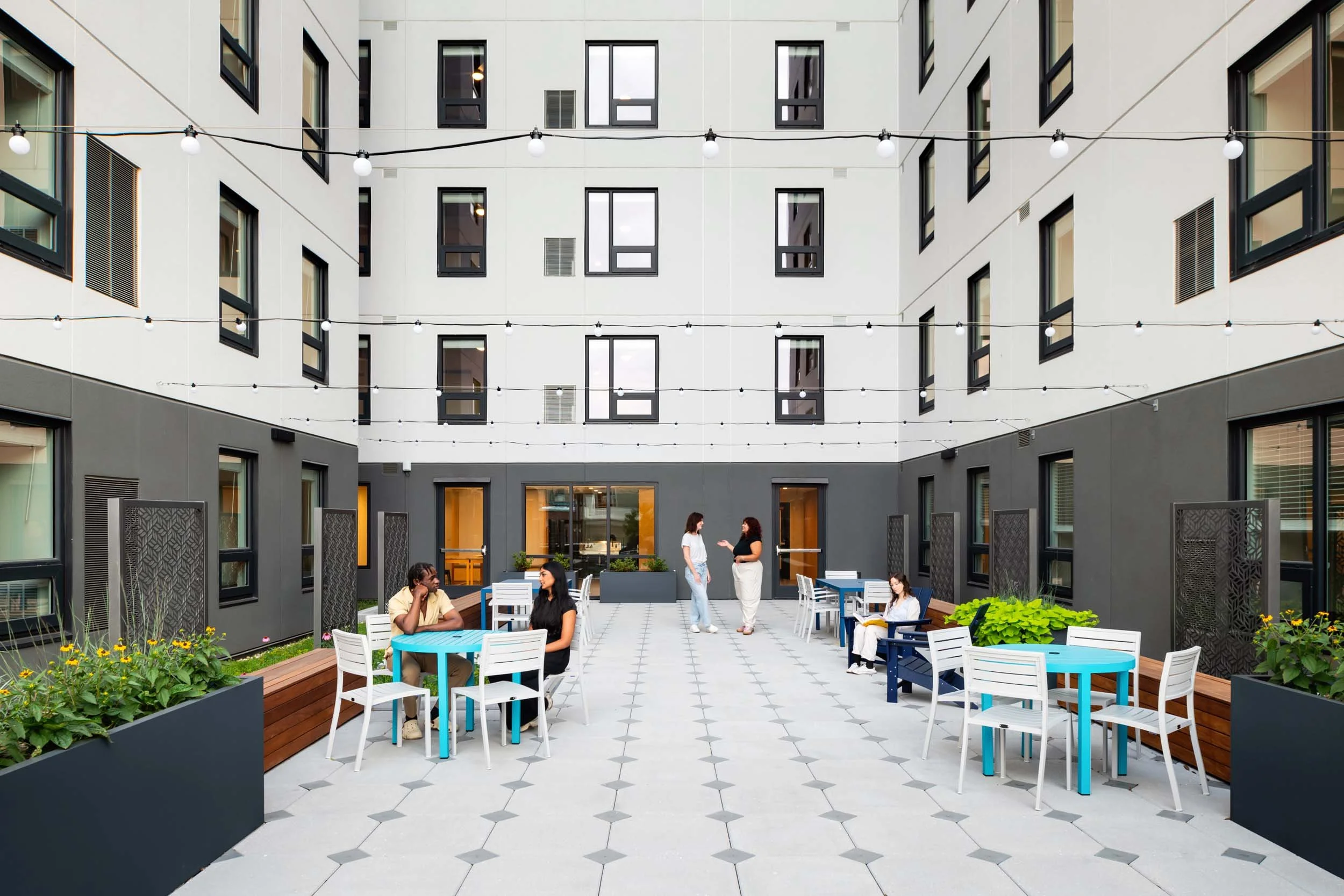 People socializing and relaxing in an outdoor courtyard of a modern apartment complex with string lights, tables, and plants.