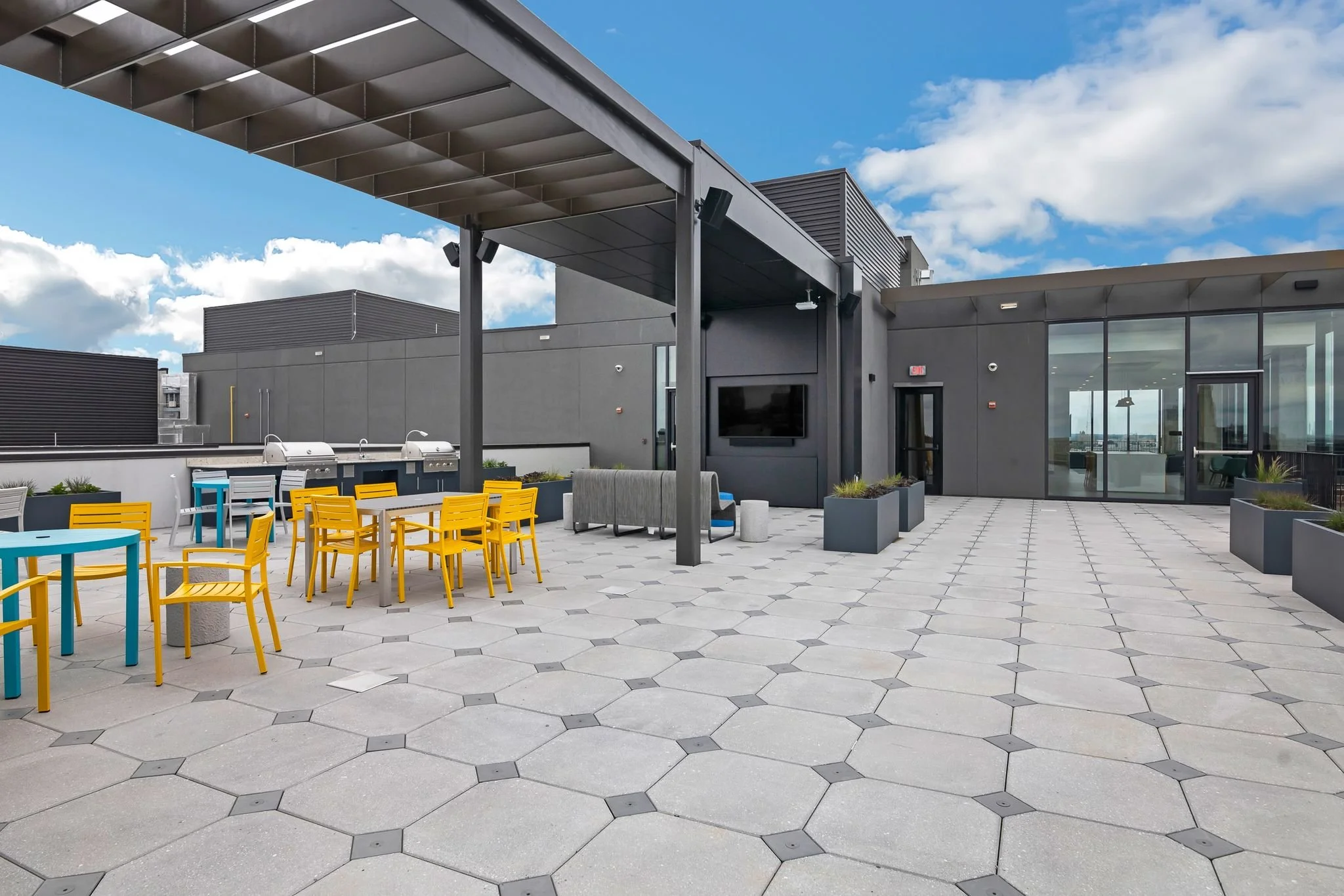 Rooftop patio with yellow and blue chairs, a grill area, and a building with large glass doors, under a blue sky with clouds.