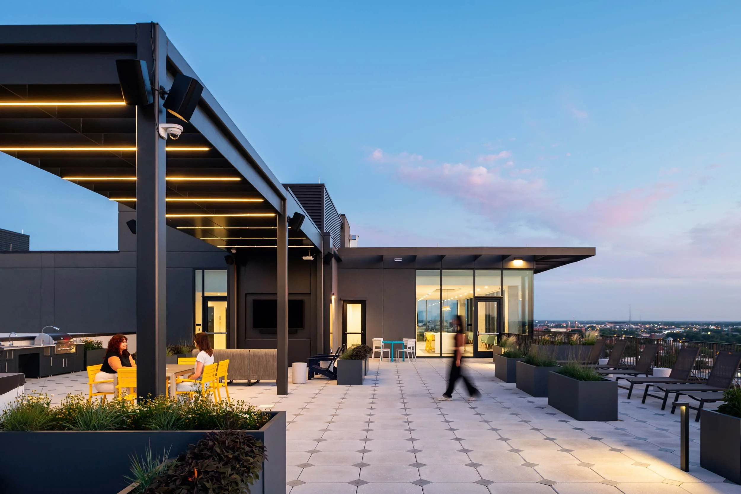 Rooftop patio with people seated at yellow chairs, modern building with large glass windows behind, city skyline in distance, evening sky with clouds, and outdoor lighting.