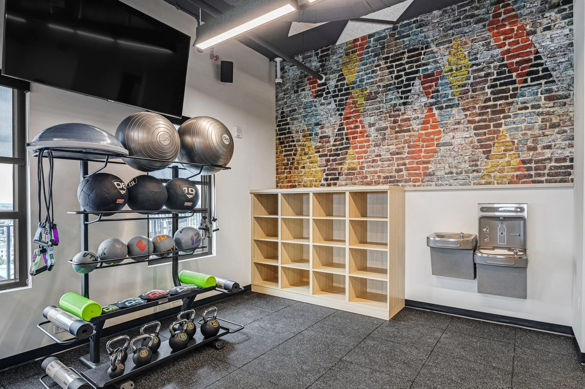 Gym corner with a black metal rack holding medicine balls, kettlebells, and foam rollers, next to a large graphic wall mural of colorful geometric faces on a brick wall, with a wooden cubby shelving unit, water fountains, and part of a window.