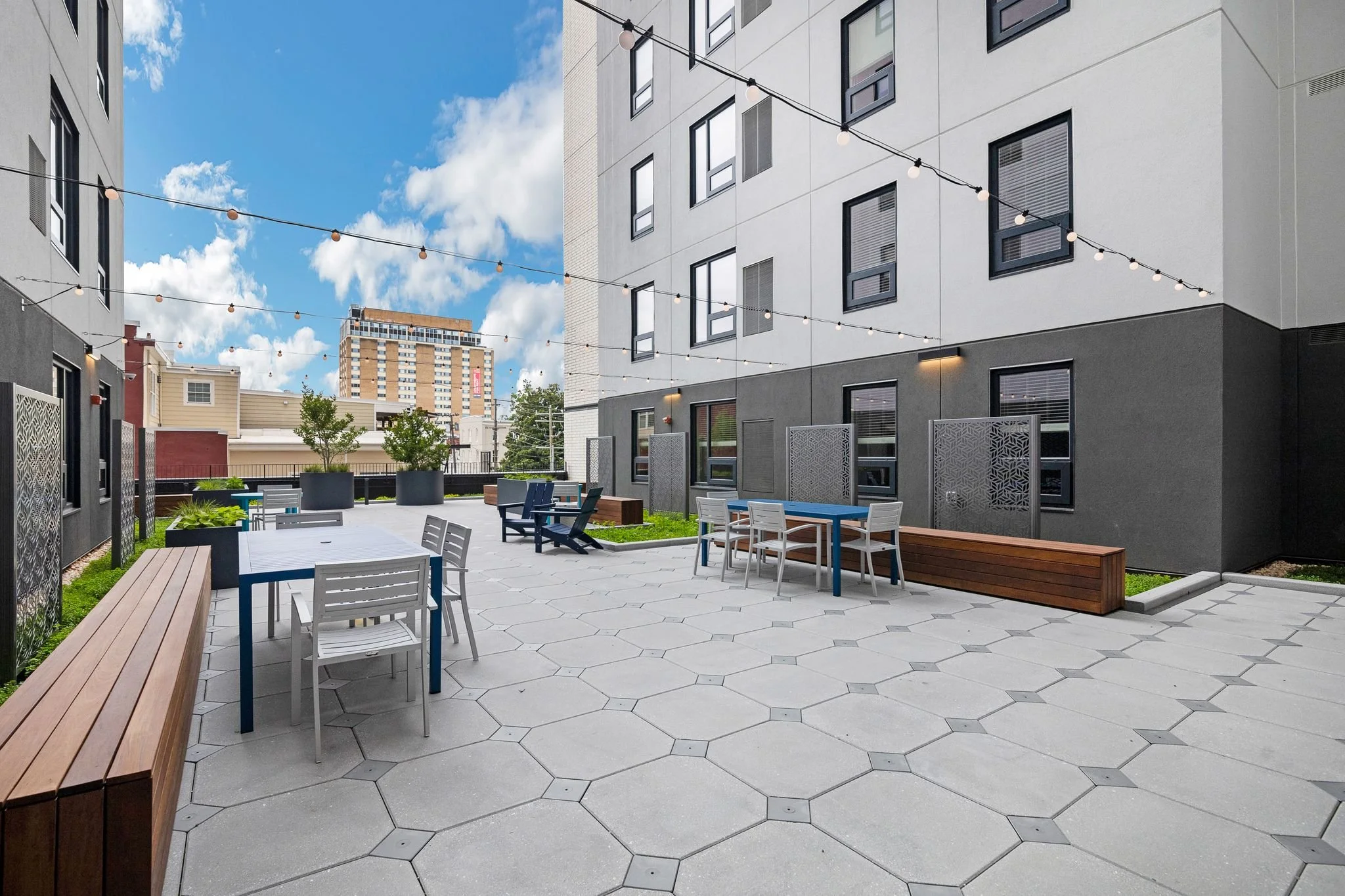 An outdoor patio area between modern apartment buildings with string lights. There are white and blue chairs around tables, potted plants, and a wooden bench. A high-rise building is visible in the background under a partly cloudy sky.