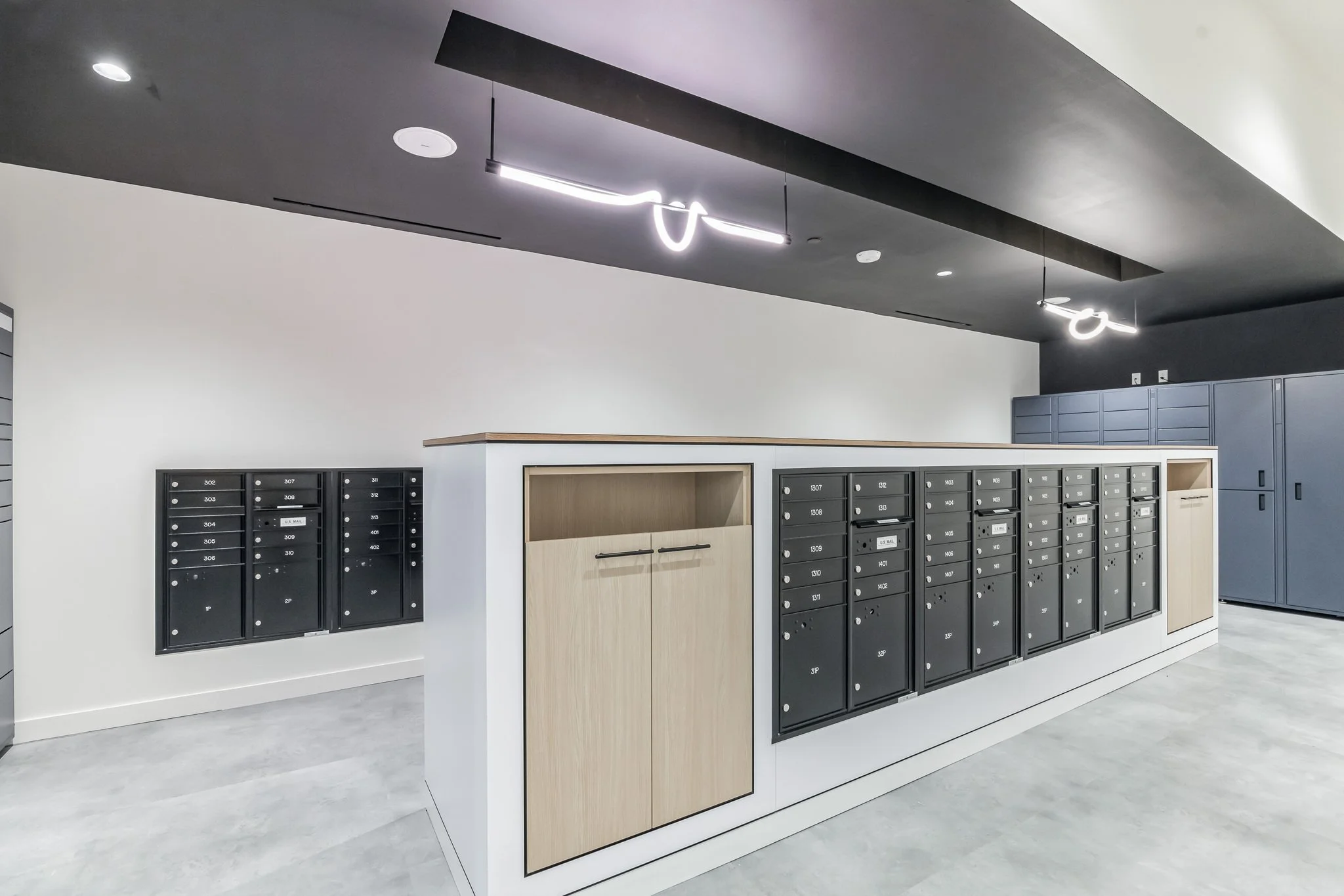 Modern mailroom with black letterboxes mounted on white wall and a white counter with a wooden accent. Fluorescent and recessed ceiling lights illuminate the space.