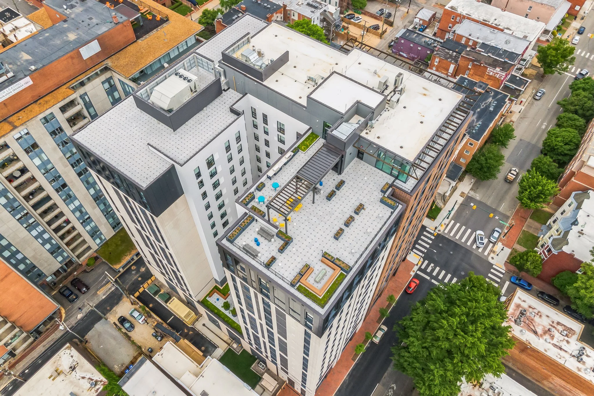 Aerial view of a modern high-rise building with a rooftop patio and surrounding city streets with trees and parked cars.