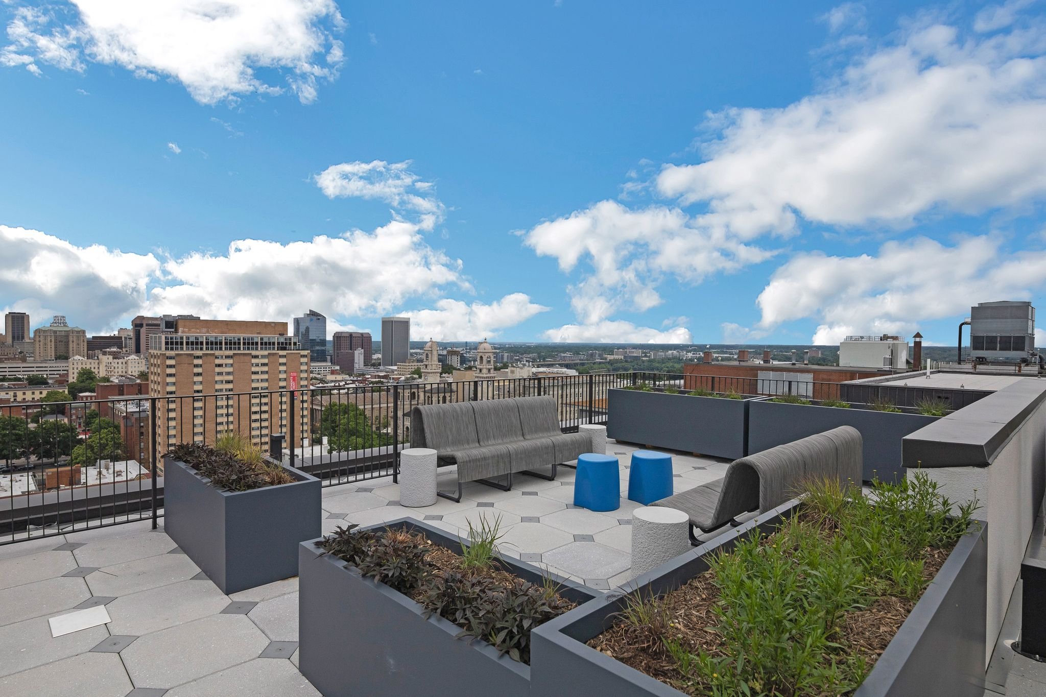 City skyline view from a rooftop terrace with modern outdoor seating, planters, and a clear blue sky with scattered clouds.