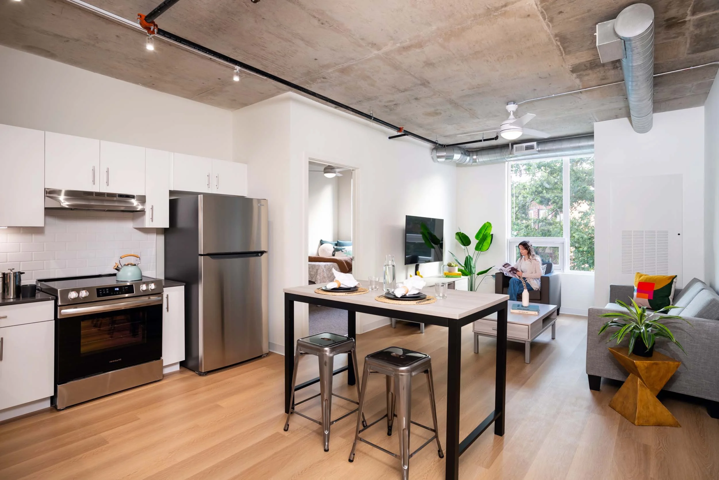Open-concept apartment with kitchen and living area, featuring white cabinets, stainless steel appliances, a black dining table with two stools, plants, and a woman sitting on a sofa reading a magazine.