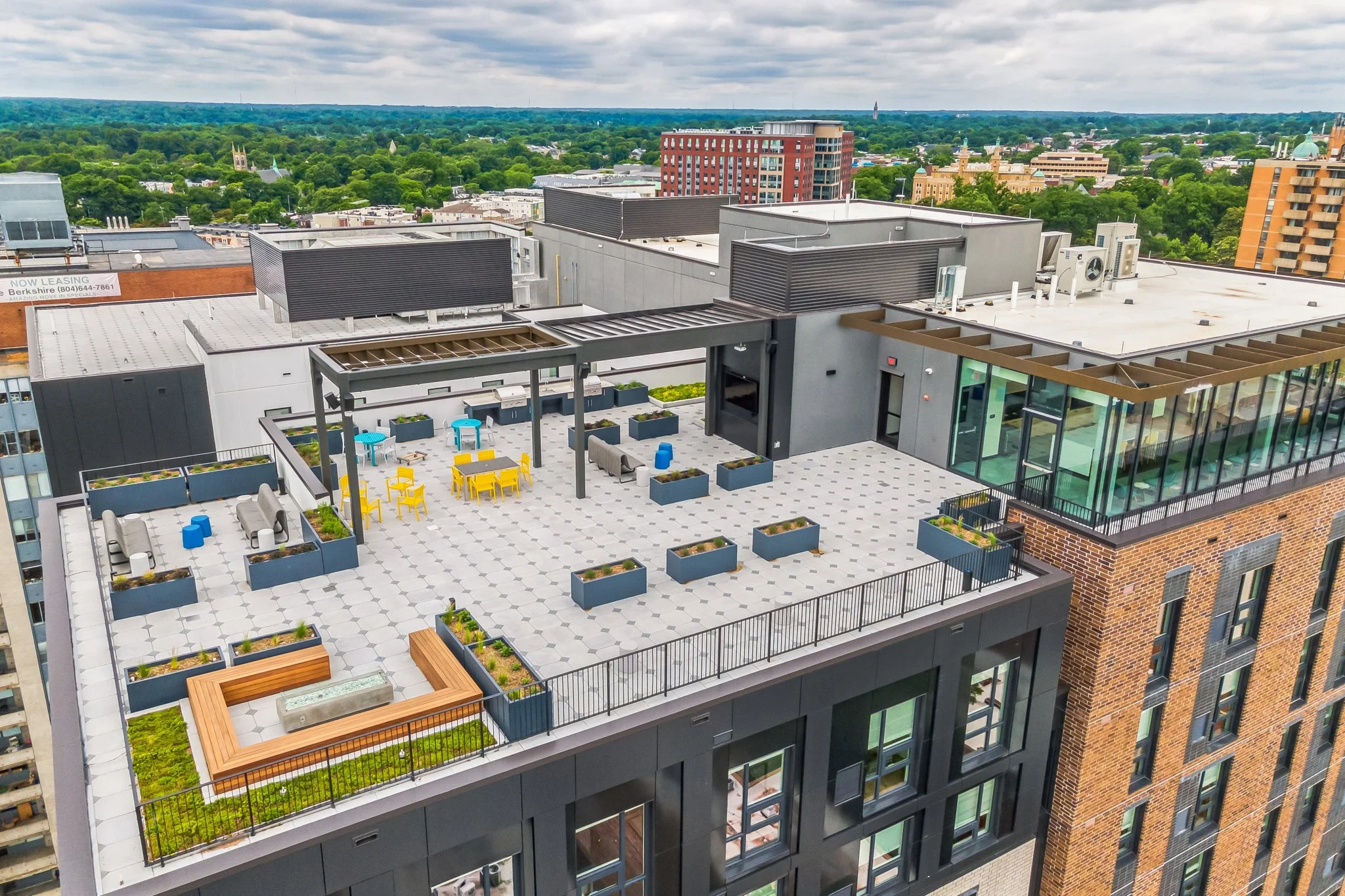 A rooftop terrace with seating and planters, overlooking a cityscape with buildings, green trees, and cloudy sky.