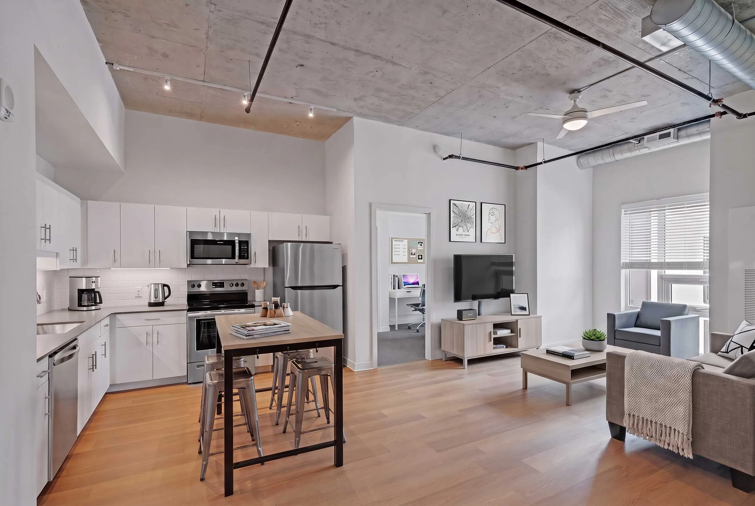Modern open-concept living space with a kitchen, dining area, and living room, featuring white cabinets, stainless steel appliances, a wooden floor, a ceiling fan, and minimalist decor.
