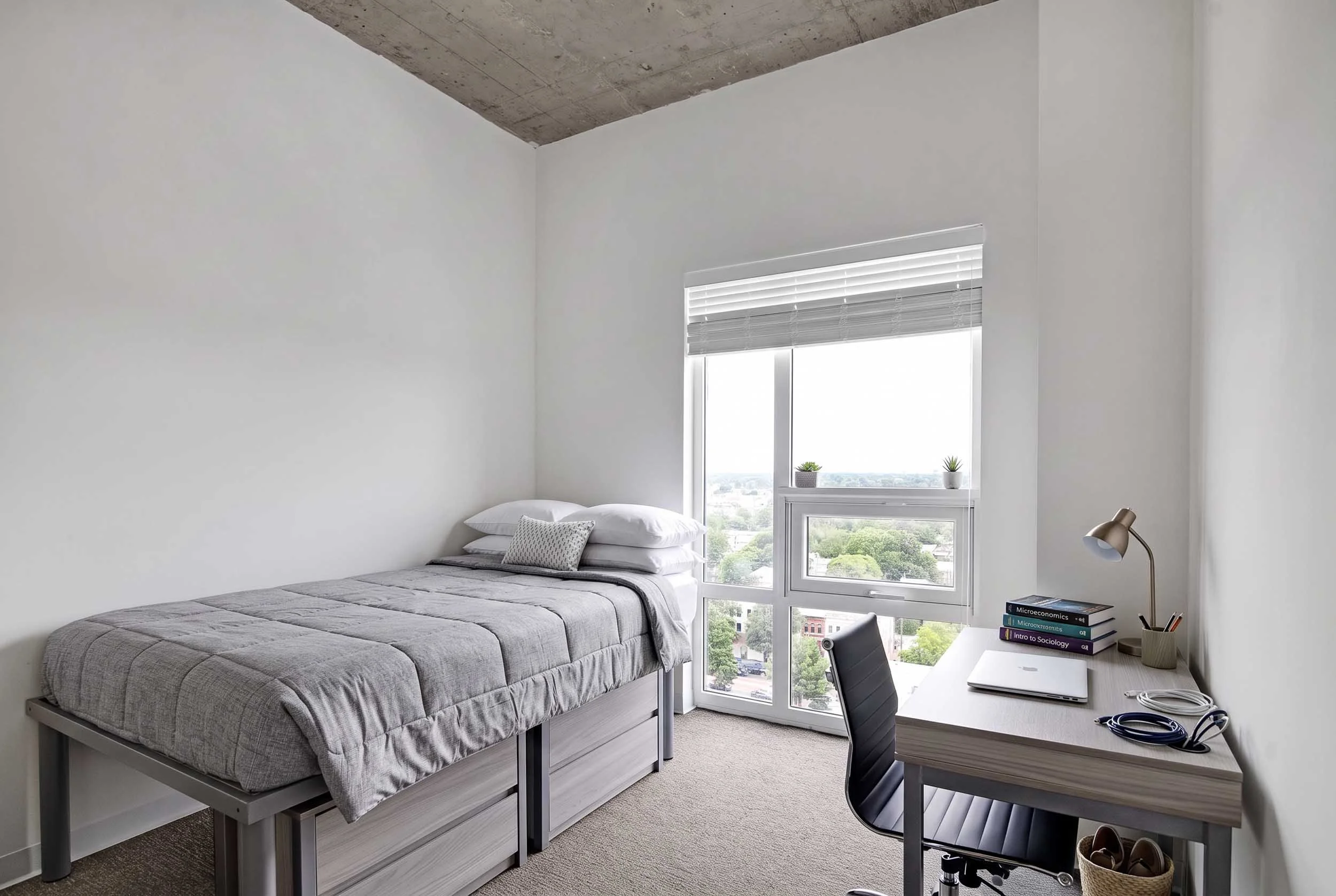 A bedroom with a bed, a window with blinds, a desk with a chair, books, a lamp, and some plants.