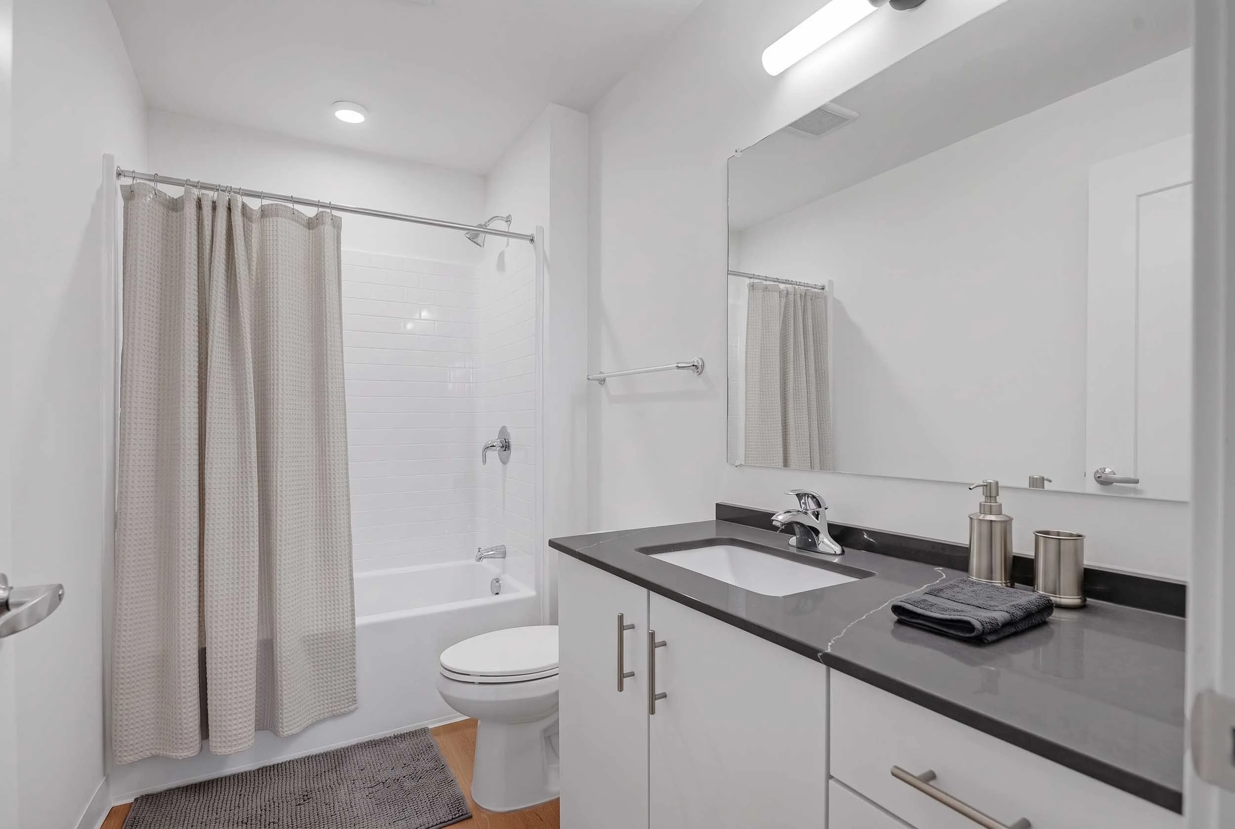 Modern bathroom with white walls, a bathtub with a shower curtain, a toilet, a black countertop with a sink, and a large mirror.