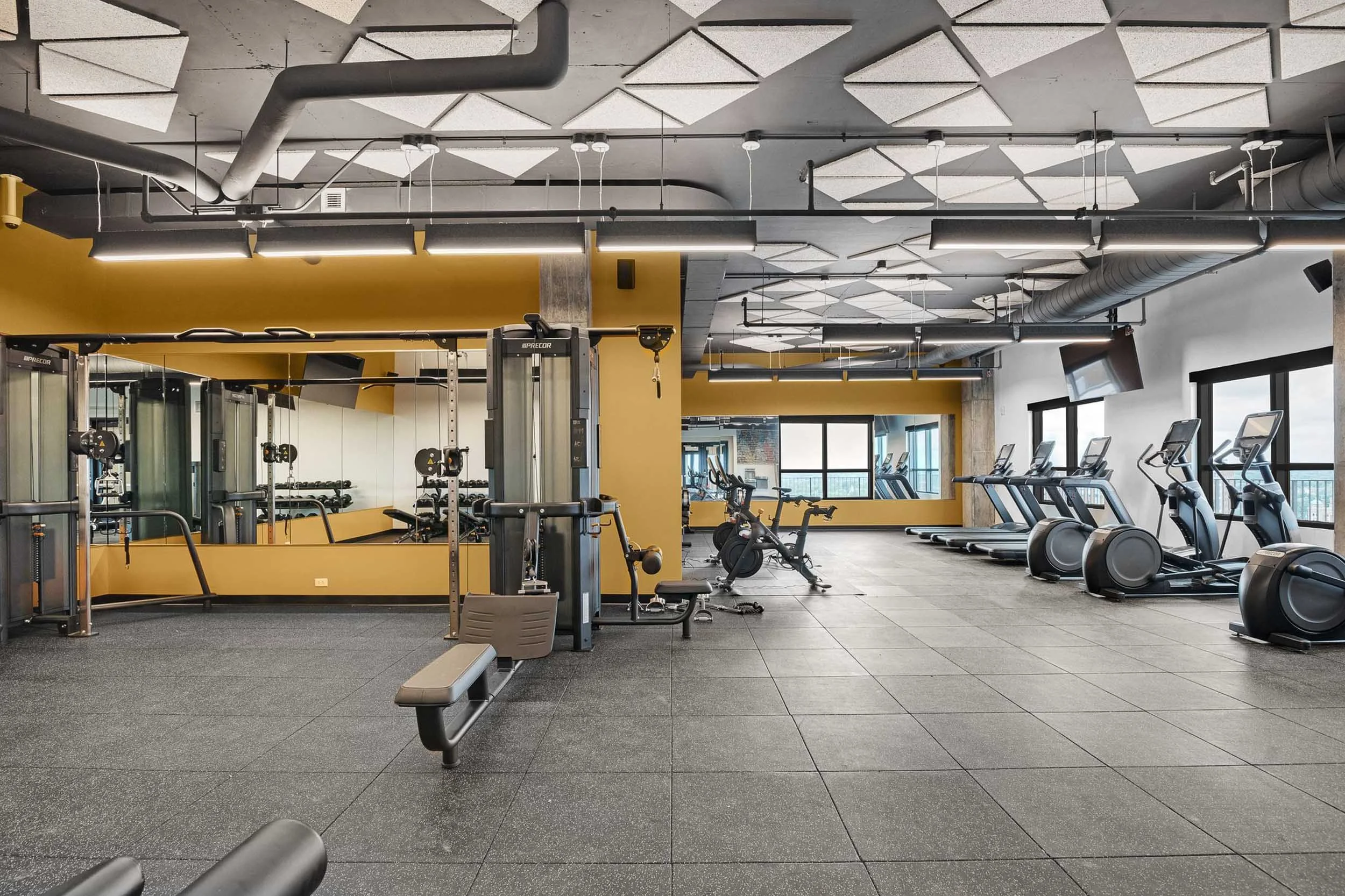 Interior of a modern gym with various exercise equipment including treadmills, ellipticals, and weight machines, with large windows and a yellow accent wall.