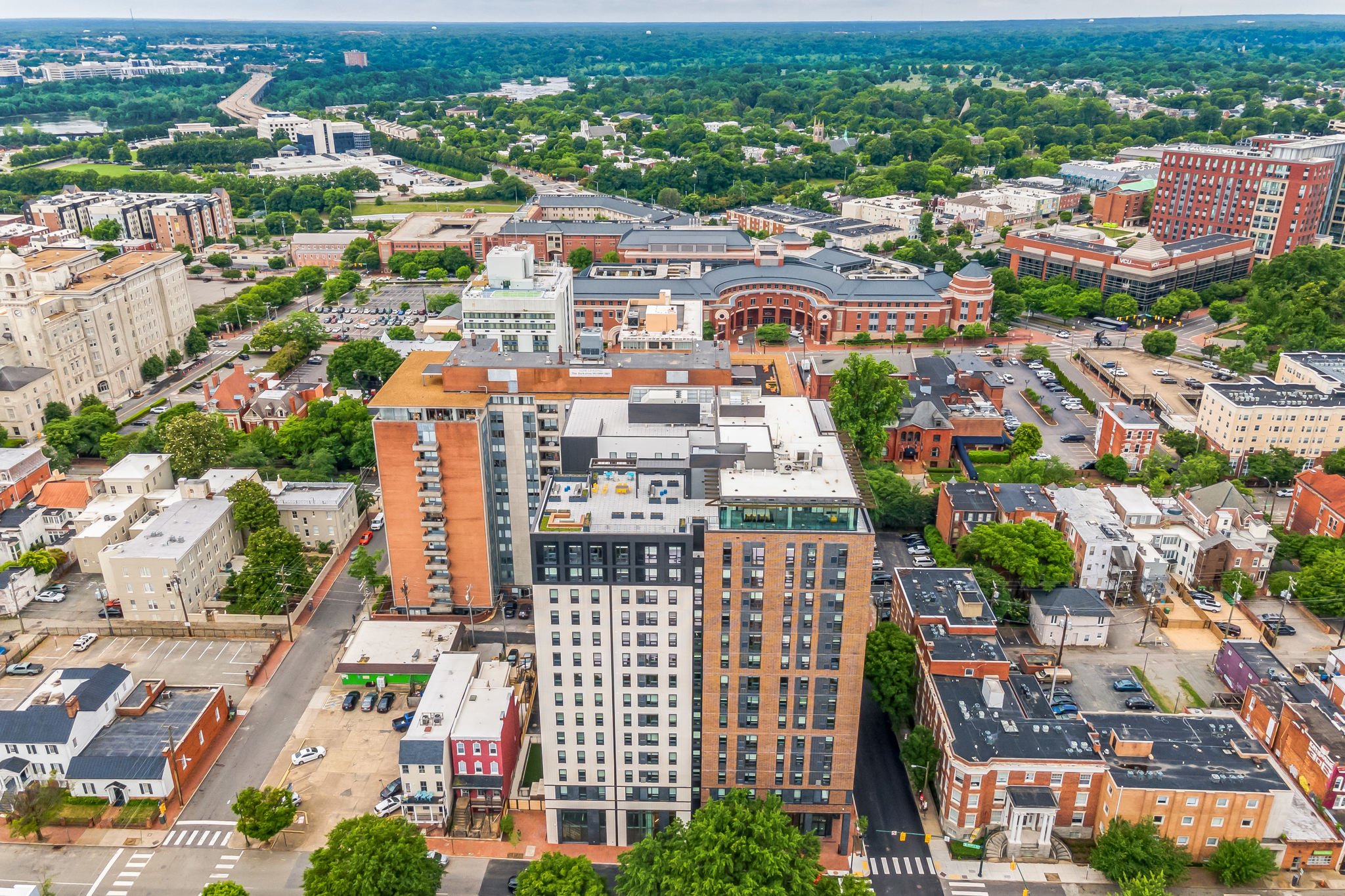 Aerial view of a cityscape with tall buildings, smaller houses, tree-lined streets, and a park in the background under a cloudy sky.