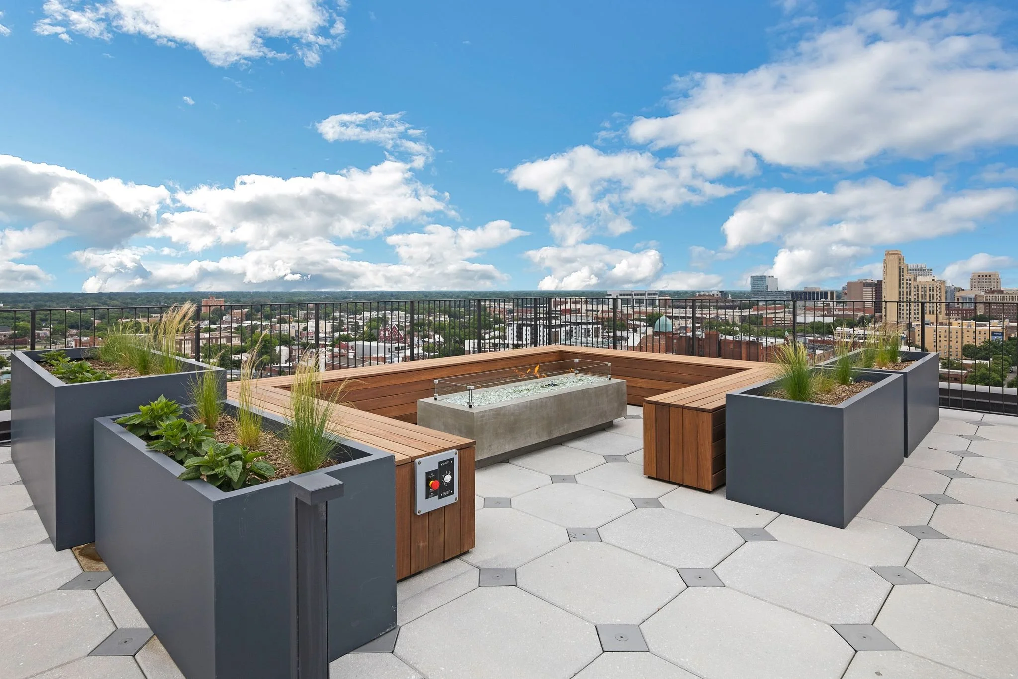 Rooftop deck with modern planters, seating, and a fire pit, overlooking a cityscape with a blue sky filled with clouds.