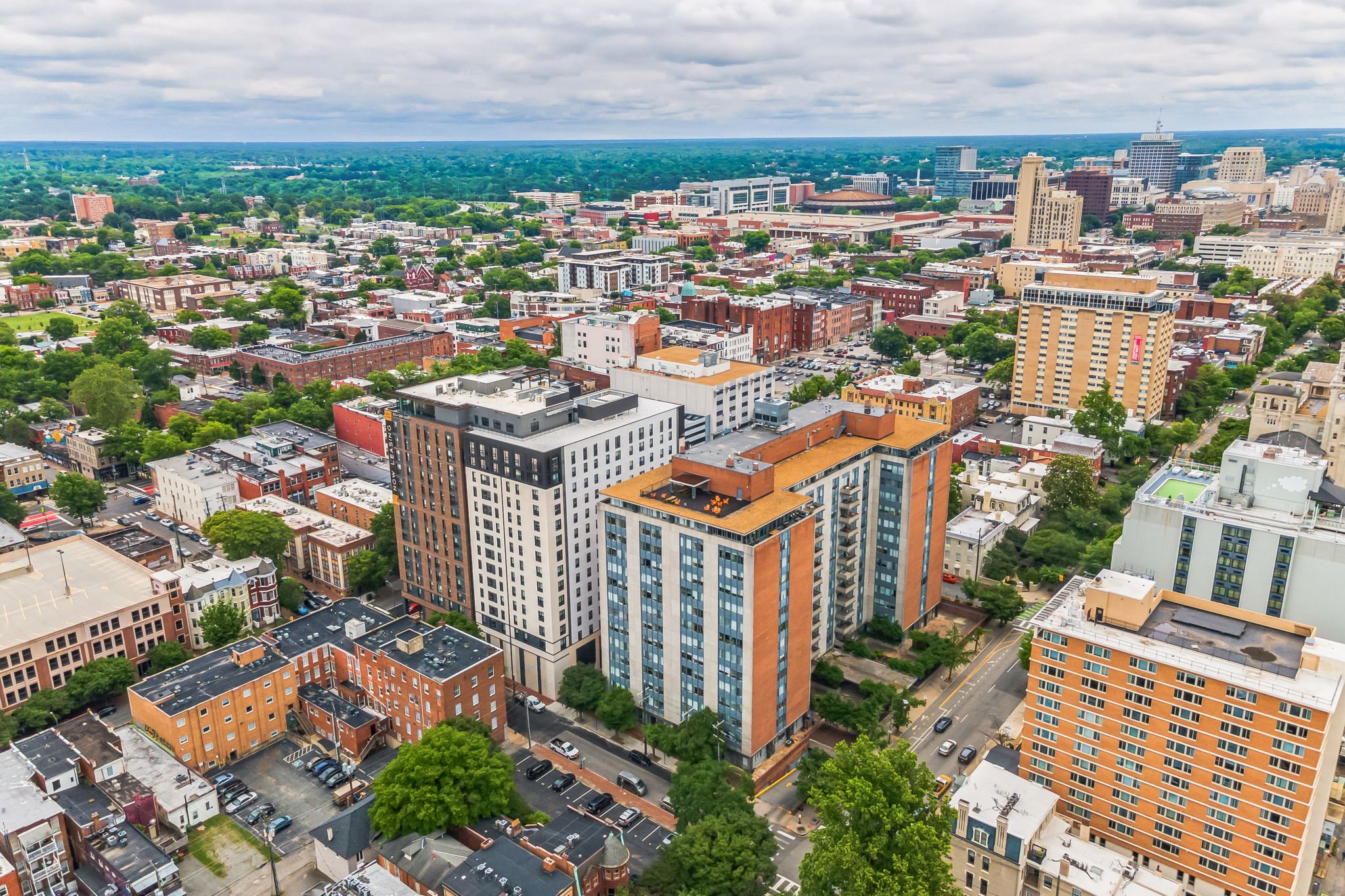 Aerial view of a cityscape with high-rise buildings, smaller shops and houses, green trees, and a cloudy sky.
