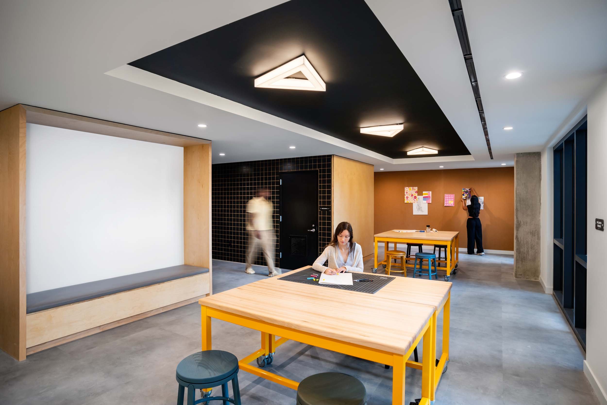 Modern indoor space with wooden tables, colorful stools, and wall art. Two women are engaged in activities; one is sitting at a table drawing, and the other is standing by a wall with artwork, while another blurred figure walks through the space.
