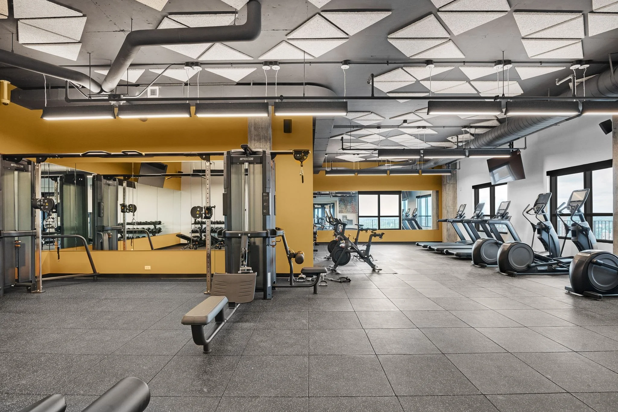 Interior of a modern gym with exercise equipment, treadmills, stationary bikes, and a weight machine. The ceiling has geometric soundproof panels and exposed ducts, with large windows letting in natural light.