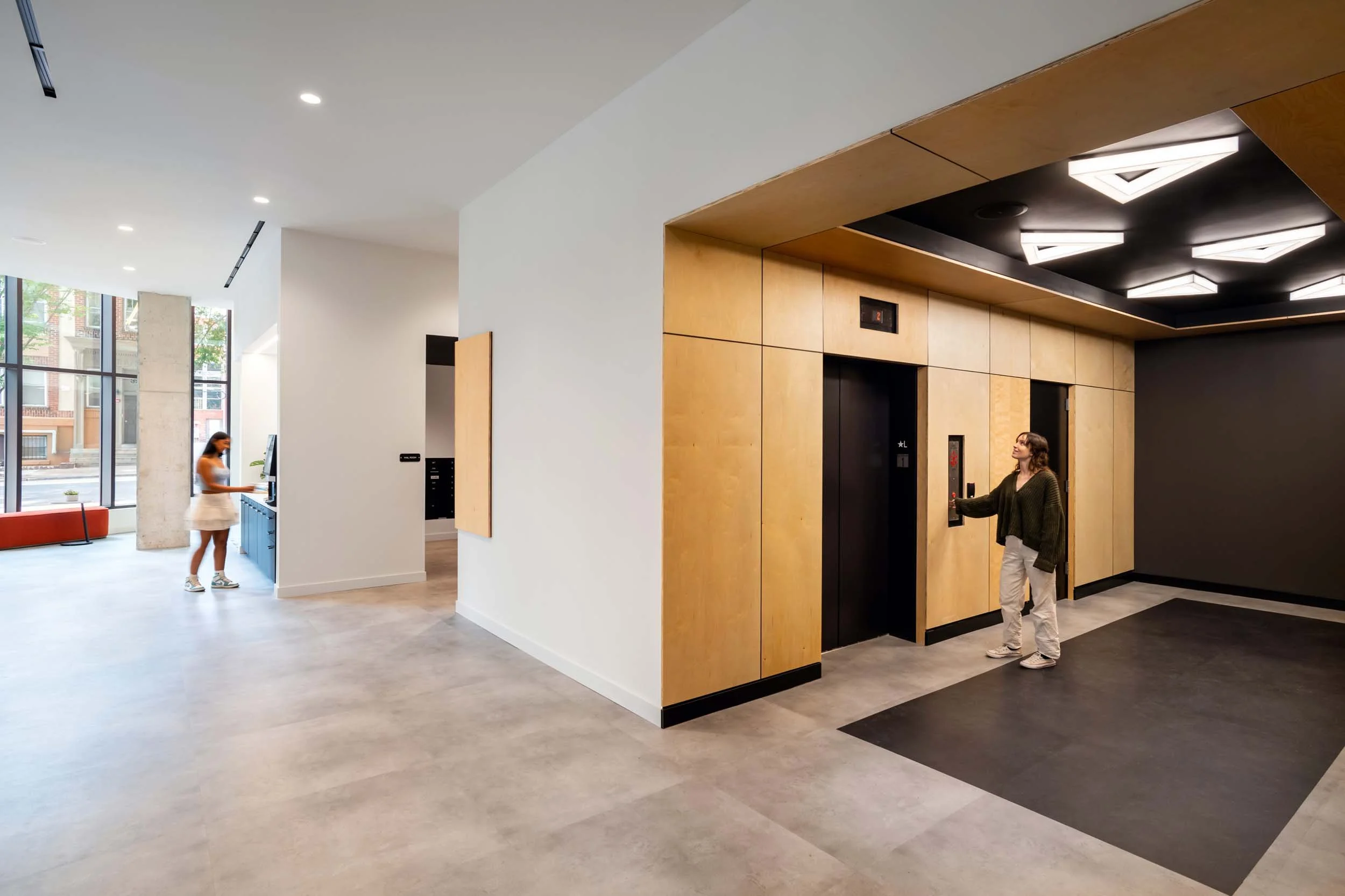 Modern lobby with elevator, two women, one using elevator and one walking. Large windows, white walls, wooden accents, geometric ceiling lights.