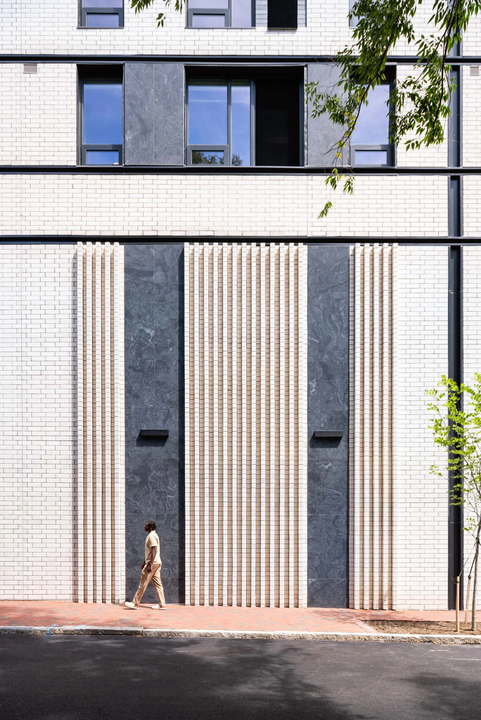 A person walking along a sidewalk in front of a modern building with a brick and marble facade, large windows, and vertical wooden slats.