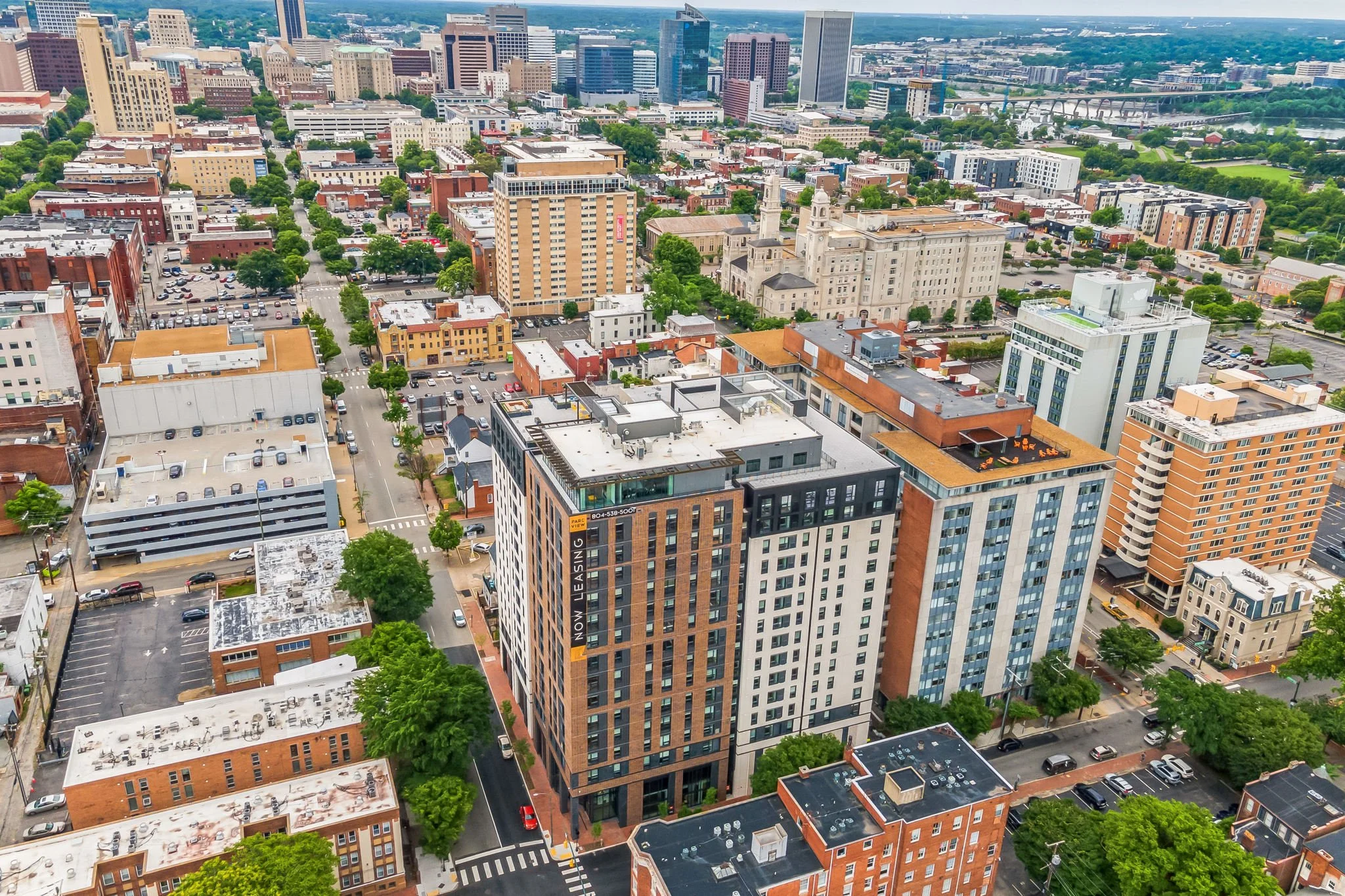 An aerial view of a cityscape featuring tall modern buildings, trees lining the streets, a large church with twin spires, and a river in the background.