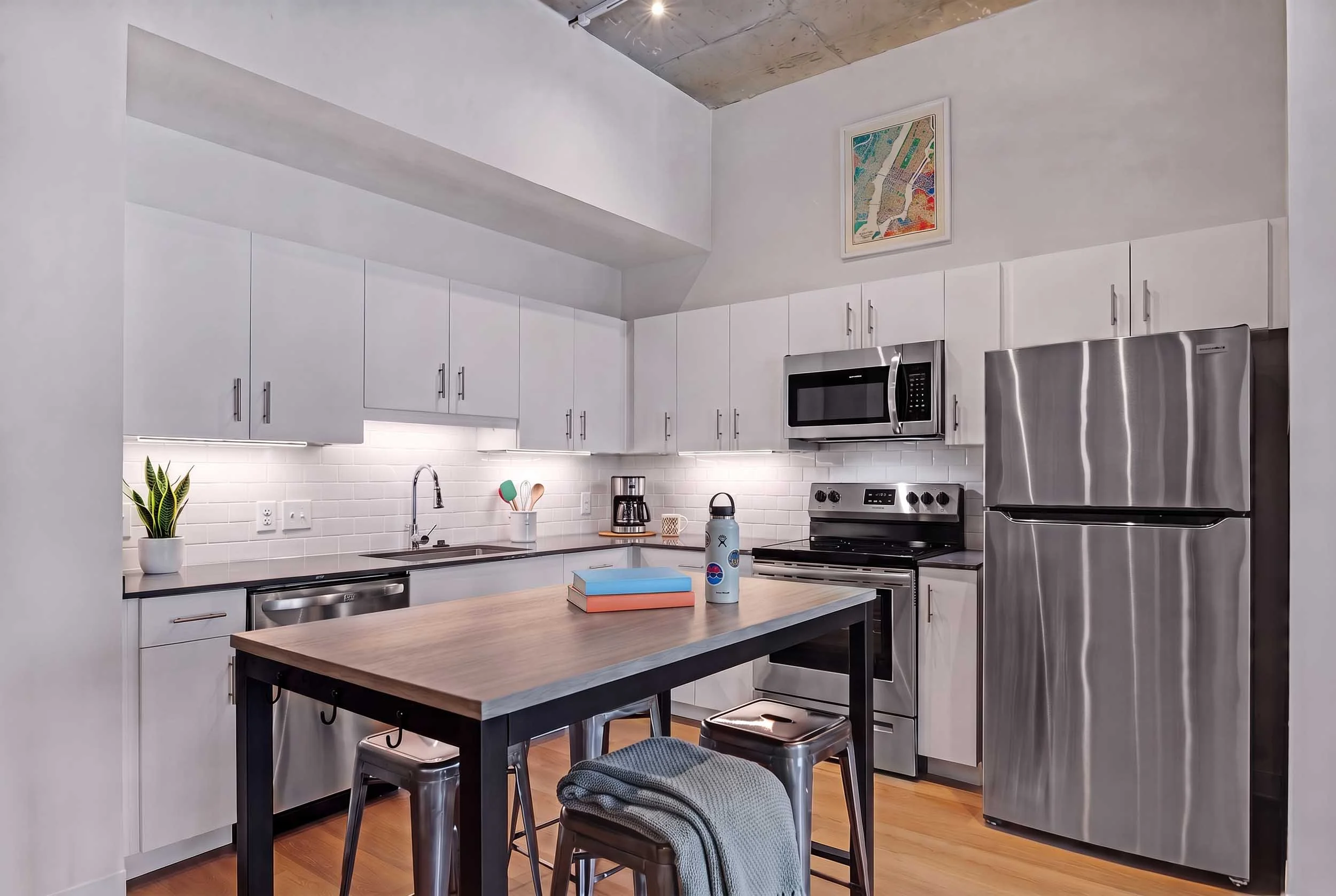 Modern kitchen with white cabinets, stainless steel appliances, a wooden island with two metal stools, a potted plant, and colorful books on the island. There is a painting on the wall and a concrete ceiling.