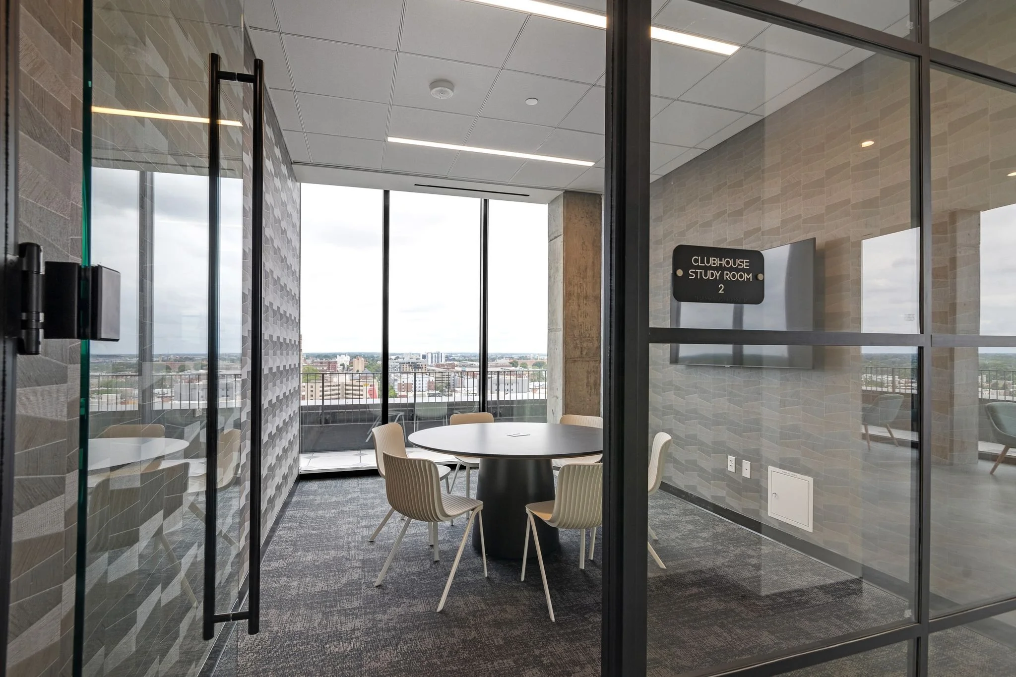 A modern conference or study room with a round table and six chairs, glass walls, and a view of the city skyline outside. There is a sign on the door that reads 'CLUBHOUSE STUDY ROOM 2'.