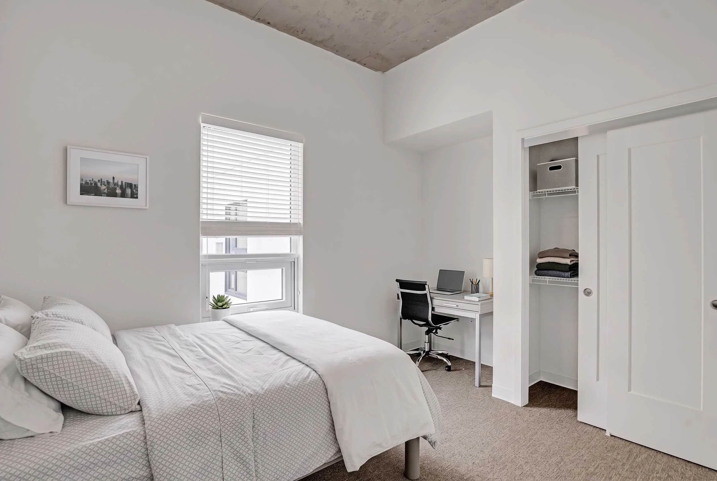 A bedroom with a white bed, a window with blinds, a small desk with a black chair, a laptop, and neatly folded towels in an open closet.