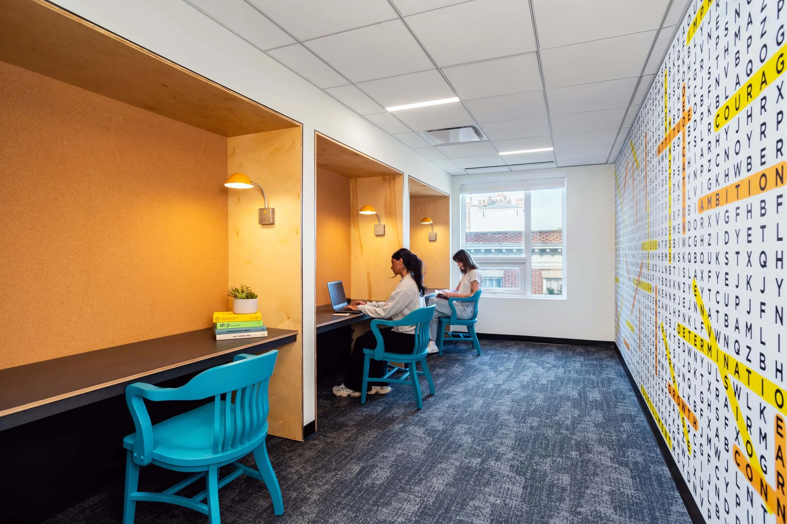 Two women sitting at desks in a modern office space with a yellow accent wall, books, potted plant, large window, and a wall with a colorful alphabet chart.