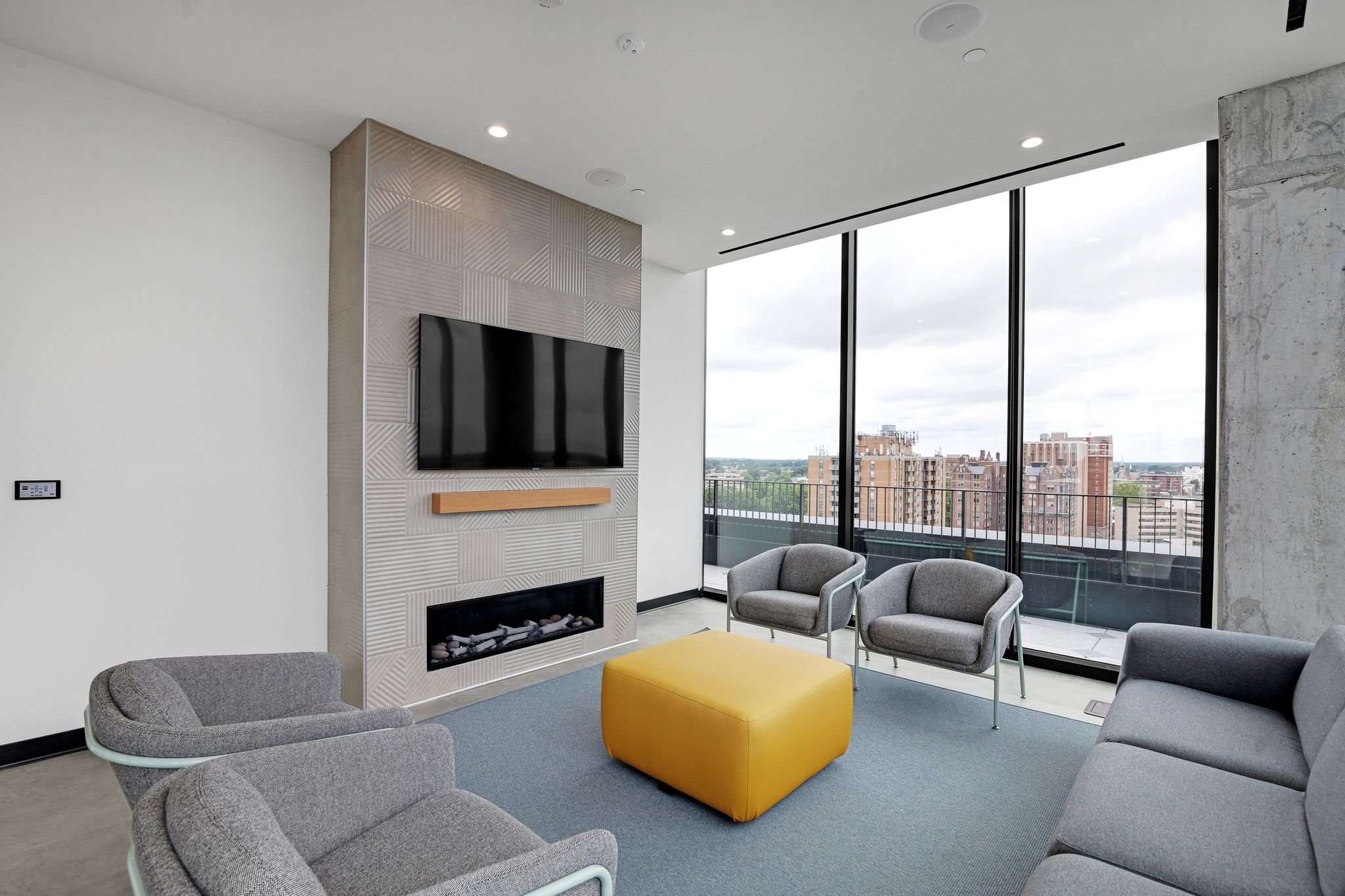 Modern living room with large windows, gray chairs, a yellow ottoman, a textured wall with a flat-screen TV, and a city view outside.