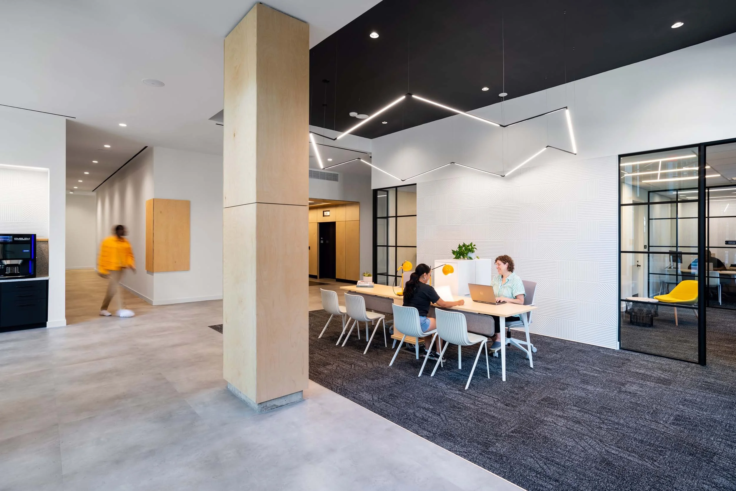 Modern office lobby with a seating area, two women working at a table, large glass partition, contemporary lighting, and a person walking in the background.