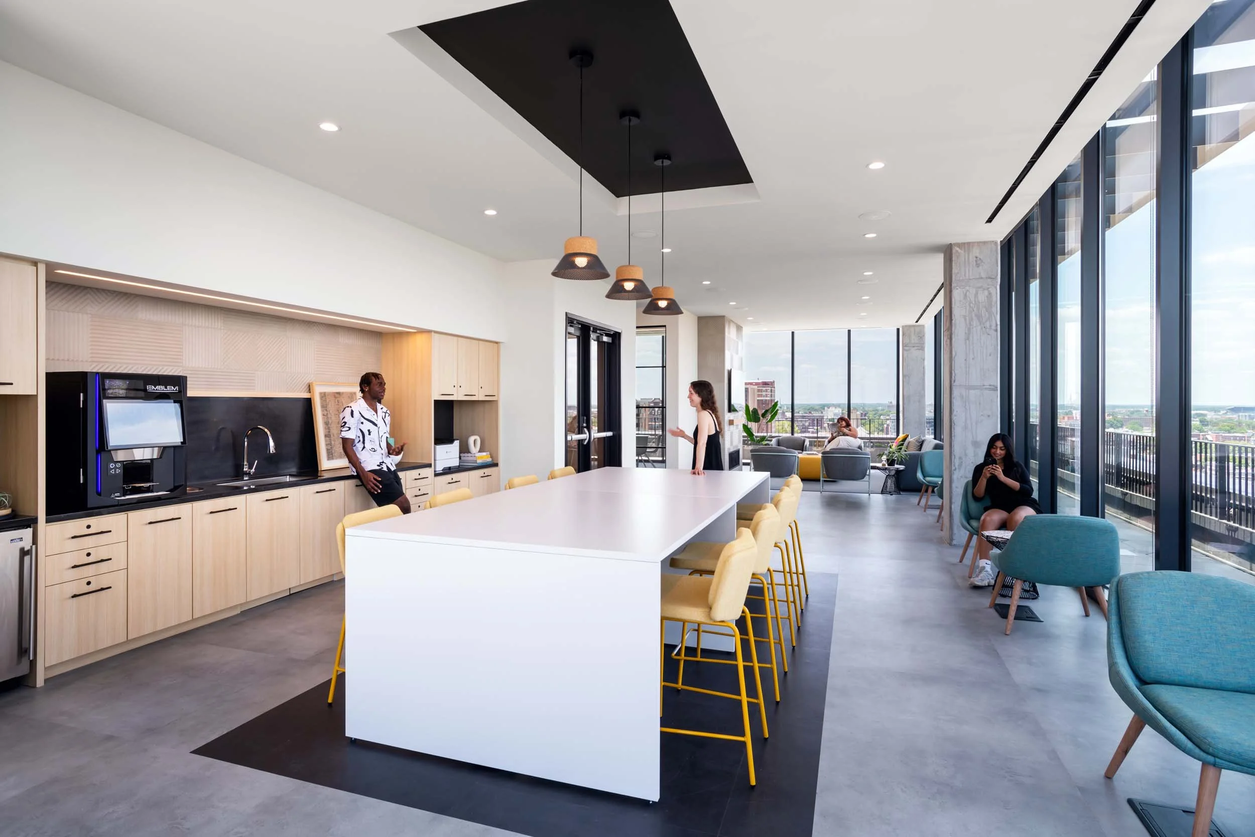 A modern lounge area with floor-to-ceiling windows overlooking a cityscape. There are several people in the space, some sitting and others standing. A long white table with yellow chairs surrounds it in the center. A kitchenette with wooden cabinets 