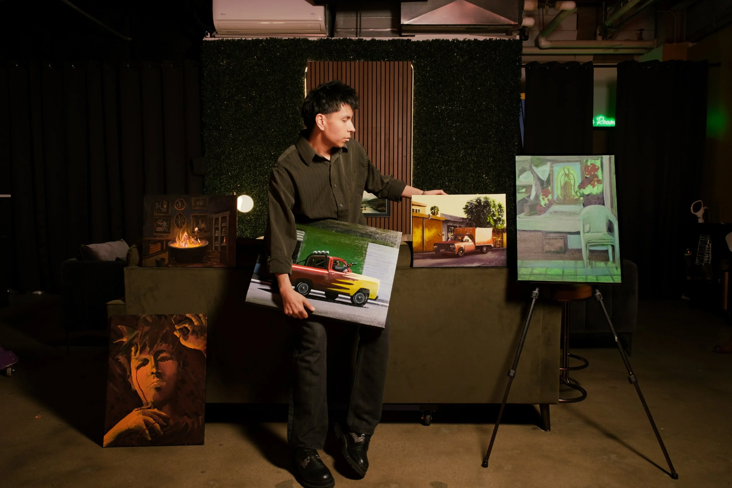Person standing in an art gallery holding paintings, with various artworks displayed behind and around them.