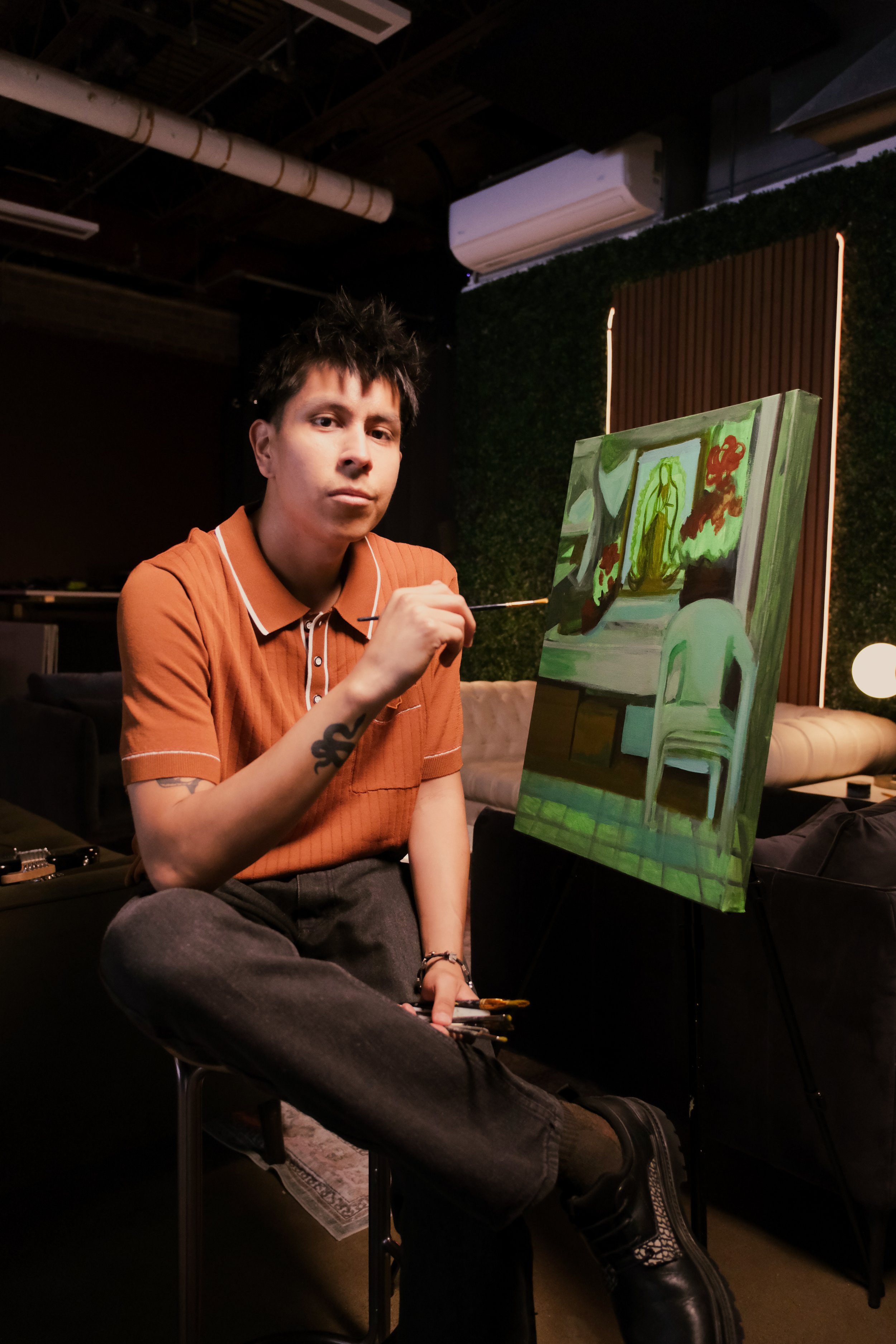 A young man with dark spiky hair, wearing an orange polo shirt, is sitting on a stool in a dimly lit room, painting a still life on a canvas mounted on an easel. The painting depicts furniture and a window with sunlight, and he is holding a paintbrush.