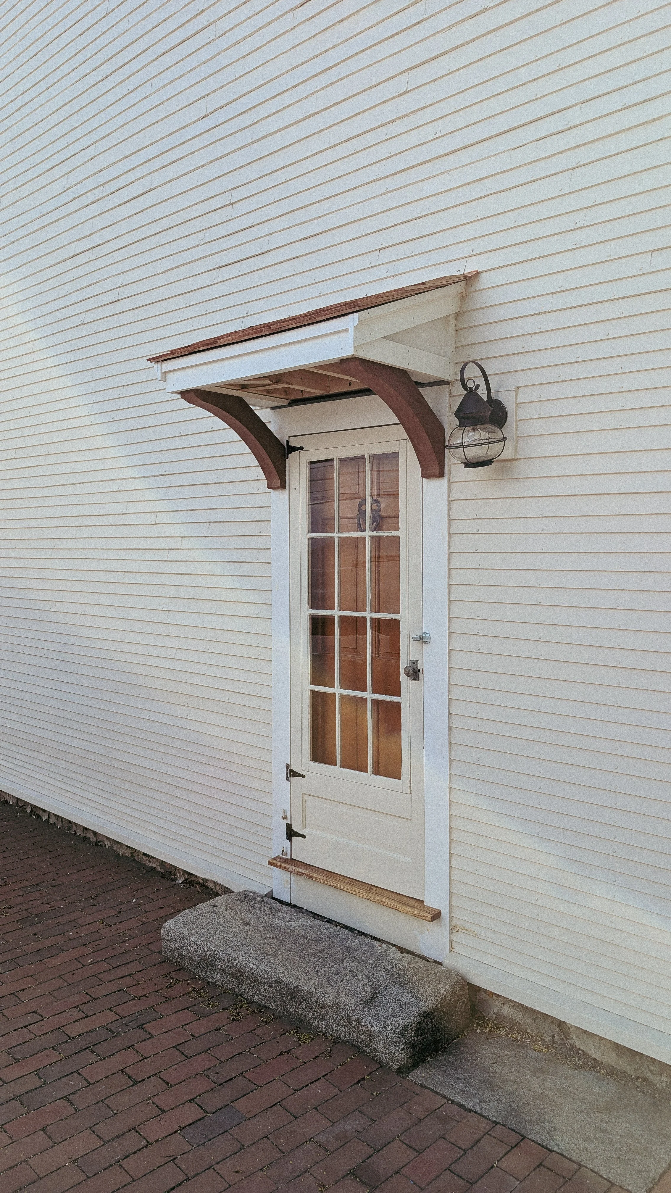 Exterior wall of a building with a white door, small awning, and a wall-mounted lantern, on a brick walkway.