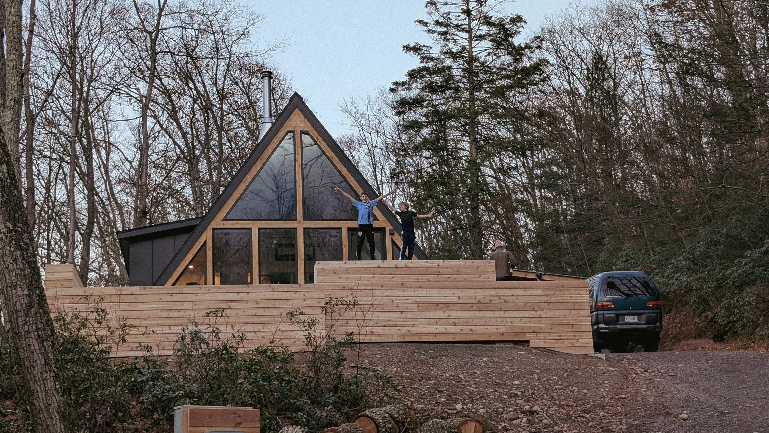 People standing on the deck of a modern A-frame house surrounded by trees during dusk, with a vehicle parked nearby.