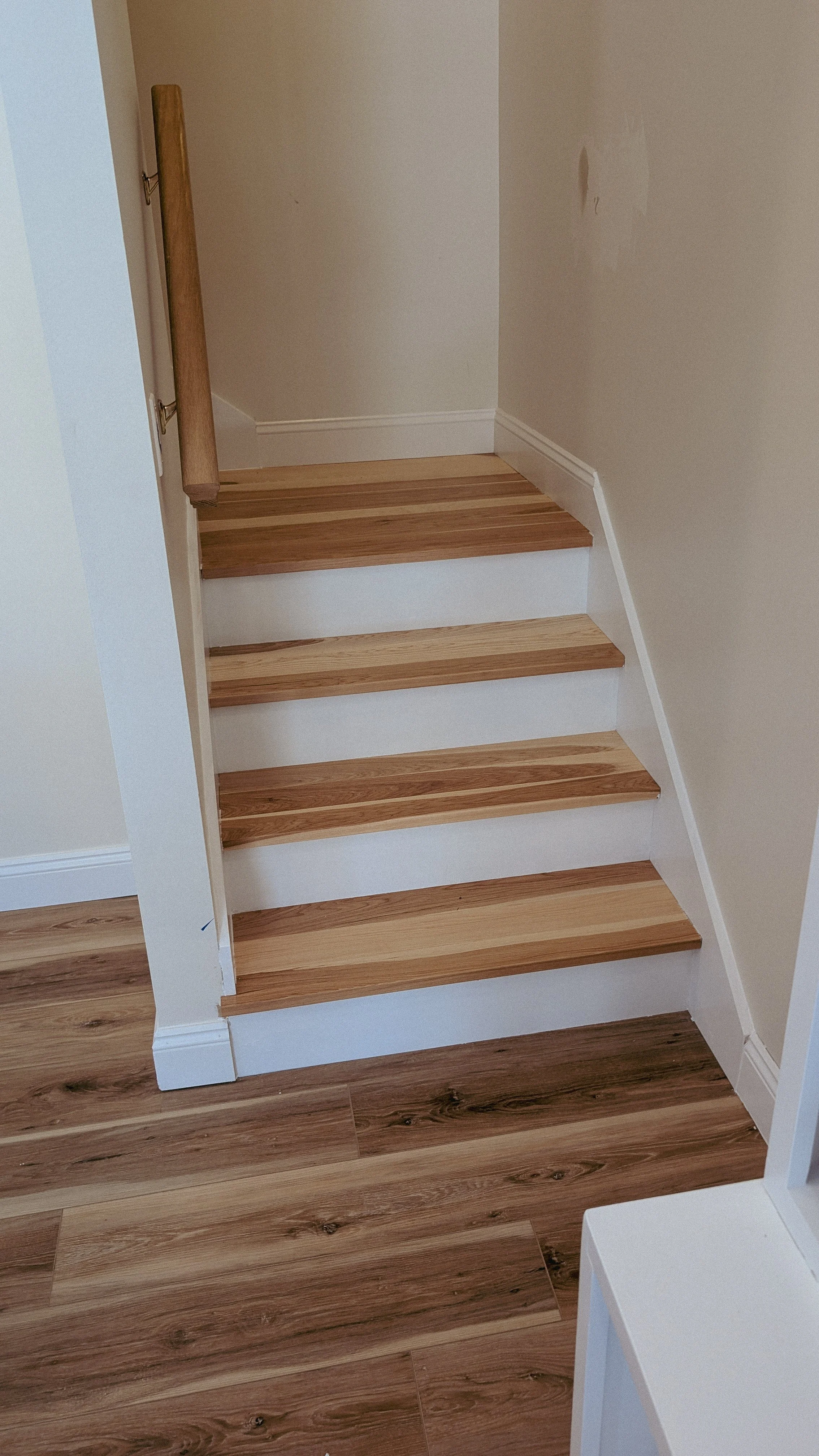 Wooden stairs with white risers in a corner, with a wooden handrail on the left side. The surrounding walls are painted a light neutral color, and a section of the wooden floor is visible at the bottom of the stairs.