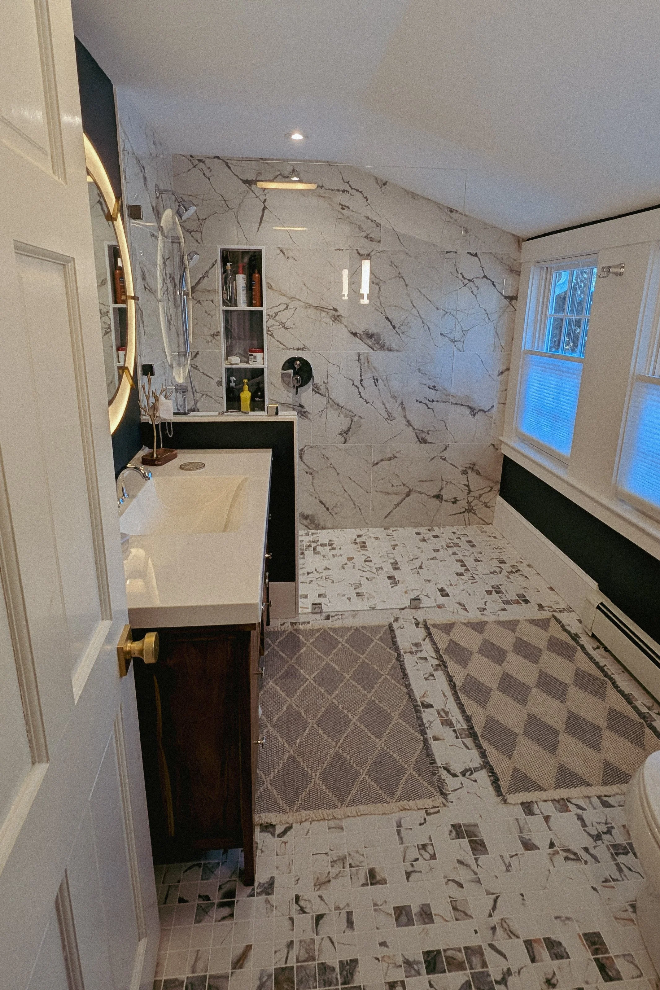 A bathroom with marble-tiled walls and floors, a window with white blinds, a white countertop with a sink, a mirror with lighting, a small cabinet with toiletries, and a patterned rug on the floor.