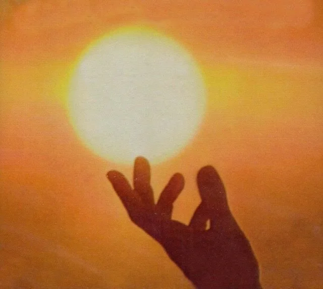A person’s hand silhouetted against a bright setting sun on the horizon.