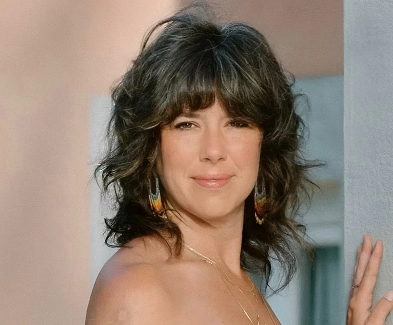 A woman with dark, curly hair and a fringe, wearing colorful earrings and a delicate necklace, standing near a wall with a slight smile.