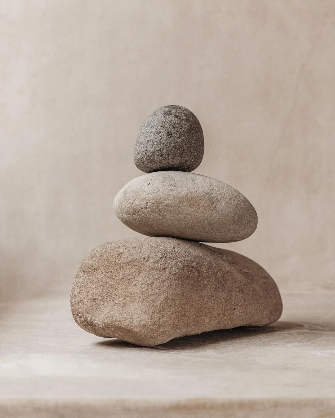 Four stacked smooth stones, varying sizes and shades of gray and beige, arranged from largest at the bottom to smallest at the top, on a neutral background.