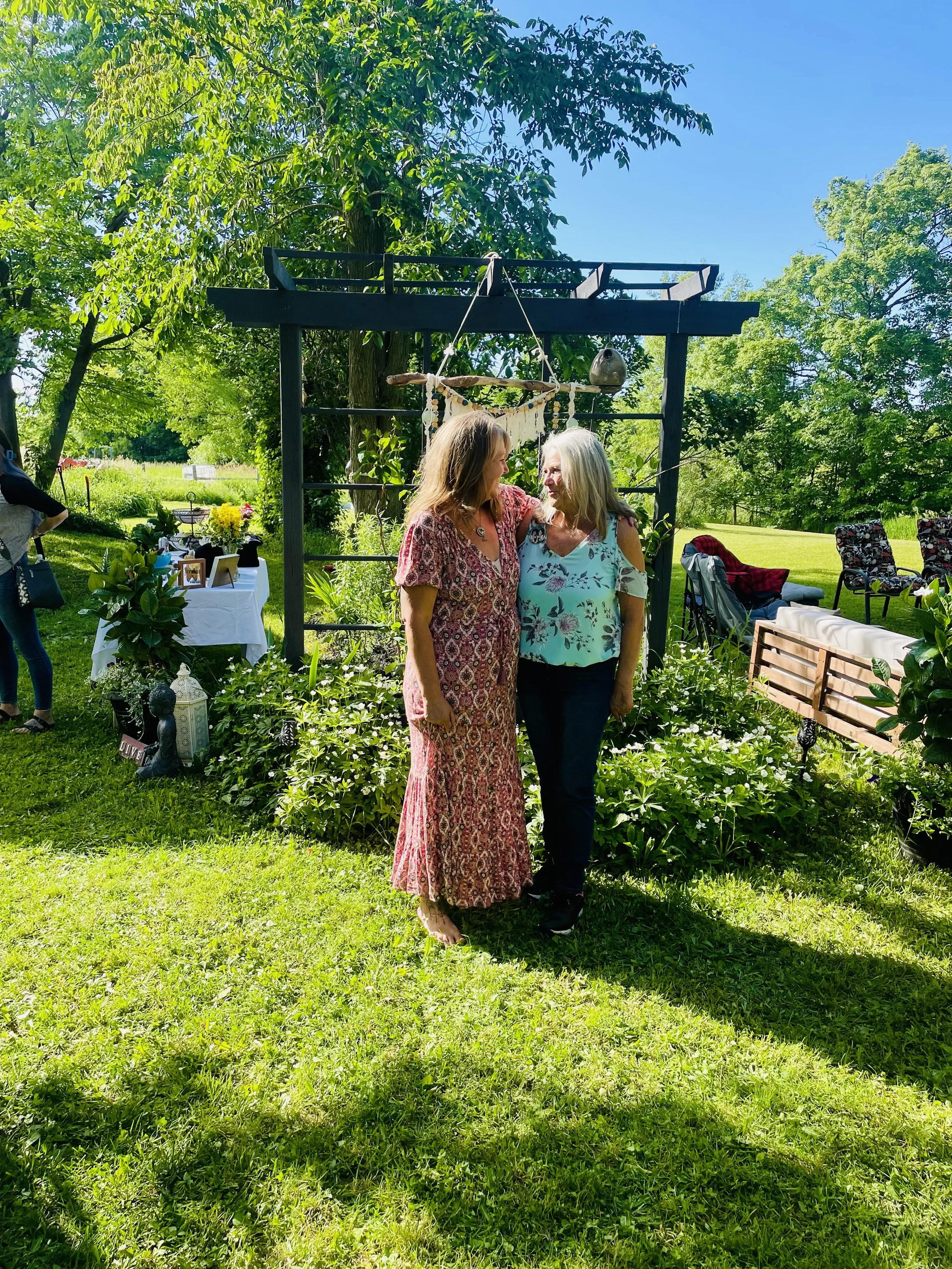 Two women are standing close together outdoors, smiling and looking at each other. They are in a lush, green garden with trees and chairs in the background, under a clear blue sky.