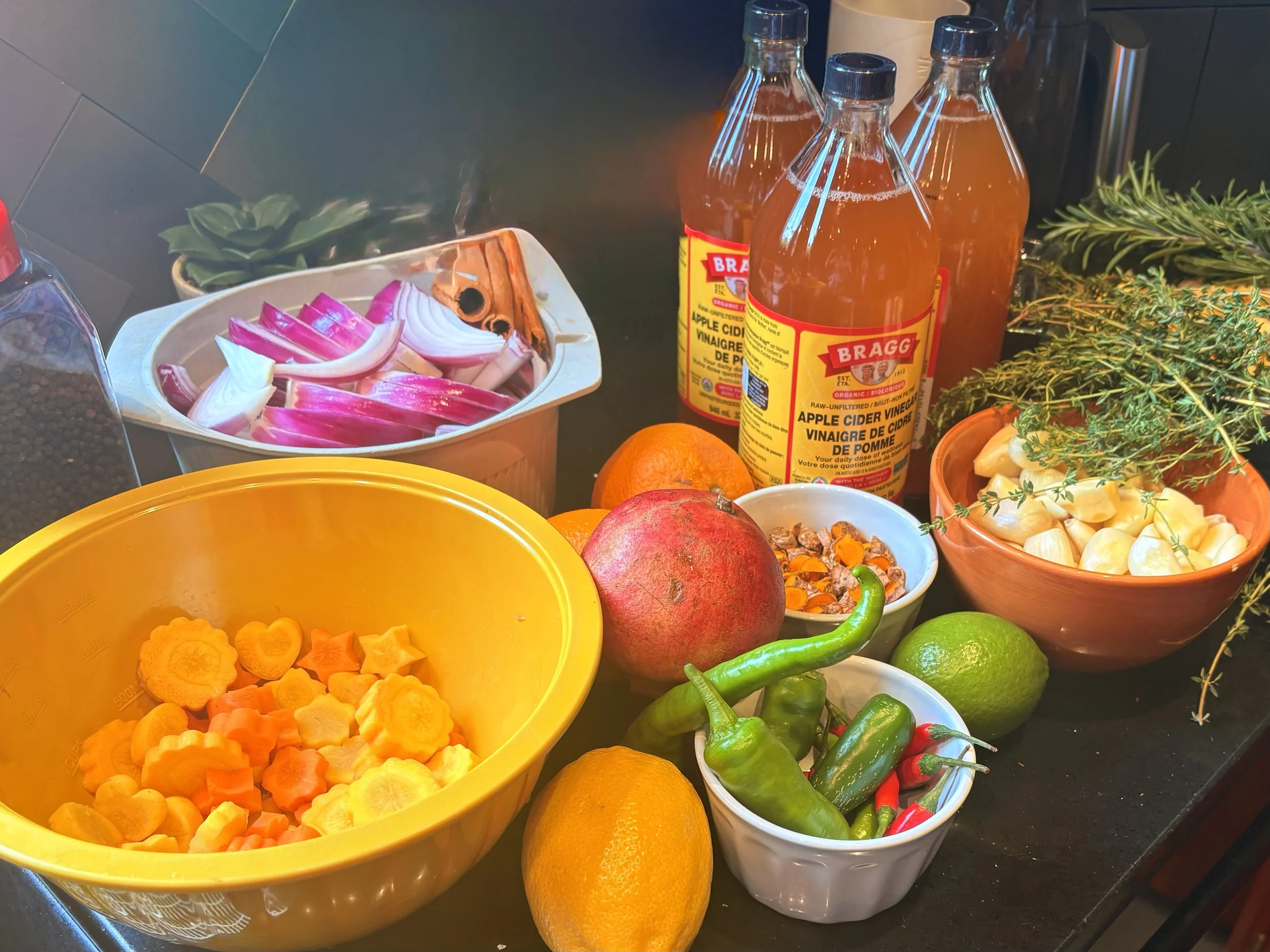 A variety of fresh produce and beverages on a kitchen counter, including sliced radishes and onions, carrots, oranges, jalapeno peppers, pomegranate, fresh herbs, and bottles of apple cider vinegar.