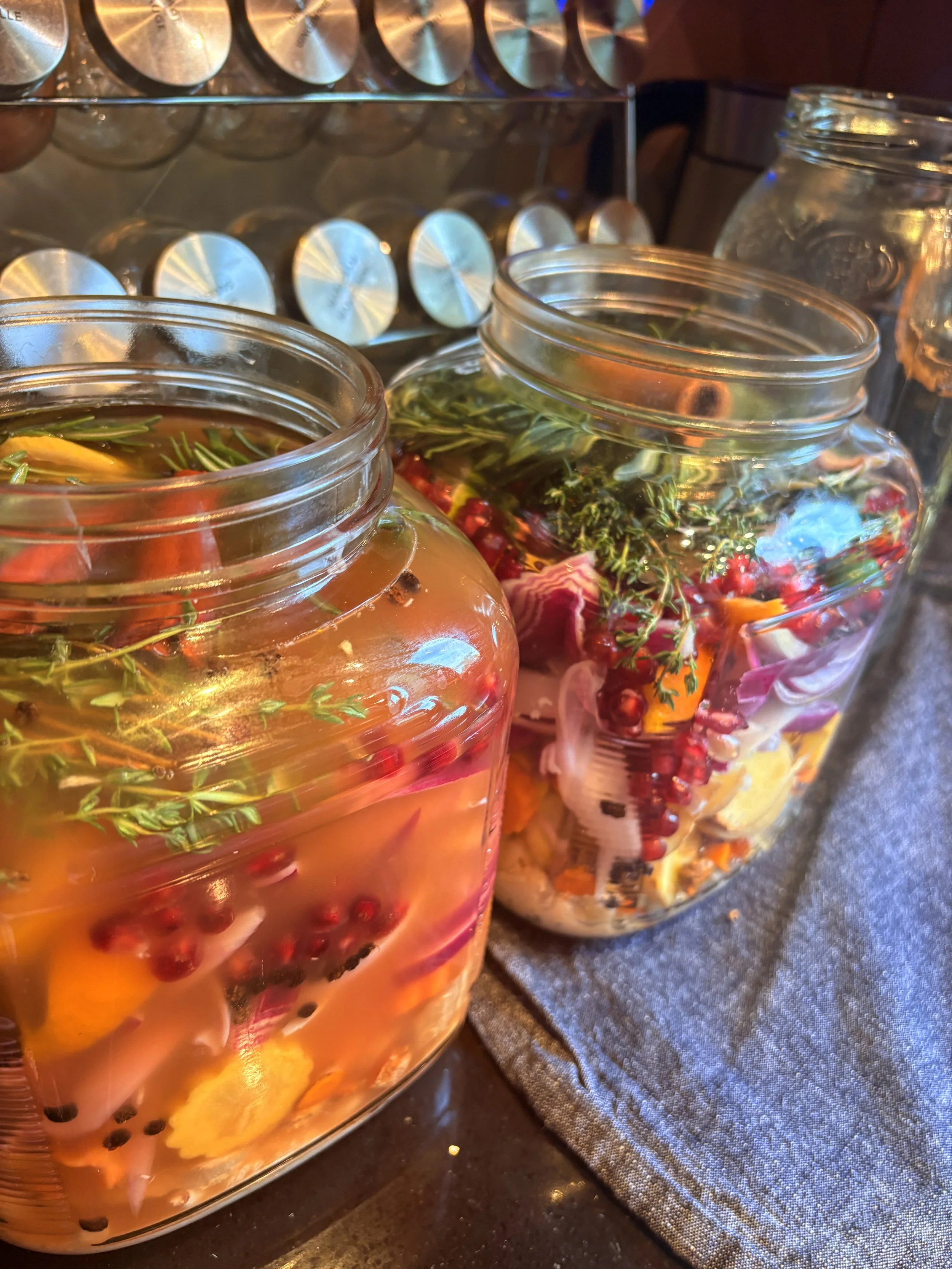 Close-up of glass jars filled with herbs, spices, and vegetables submerged in liquid, with a metal rack holding aluminum cans in the background.