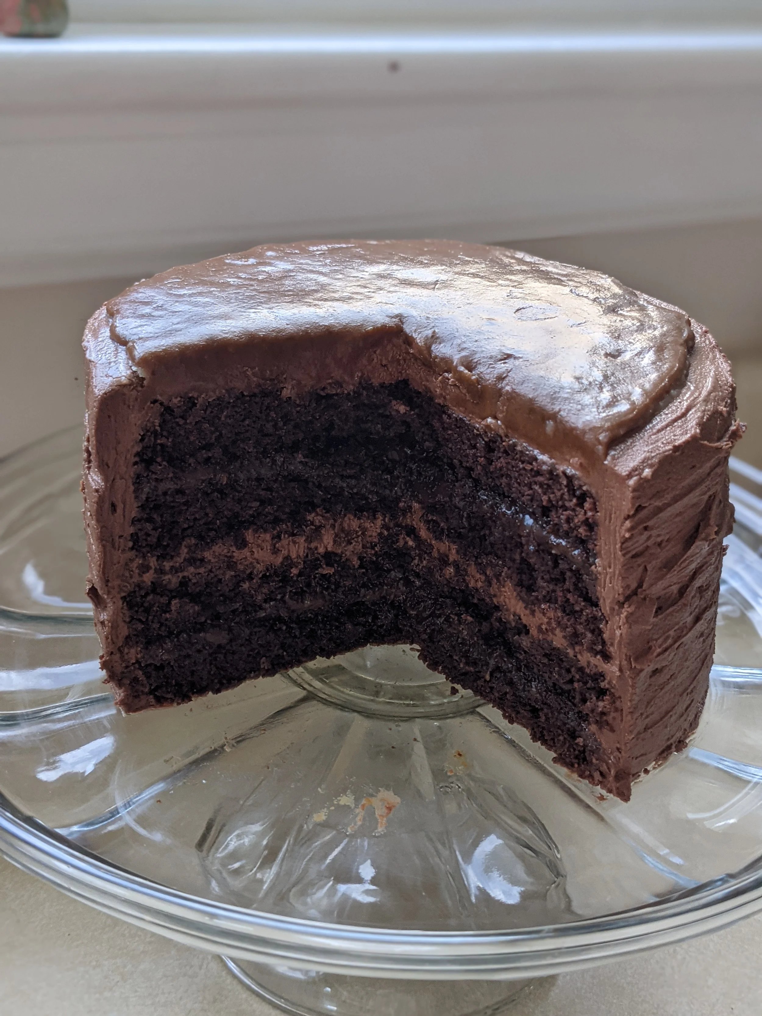 Three-layer rich chocolate fudge cake sliced open on a glass cake stand in natural window daylight, showcasing crumb texture and filling.