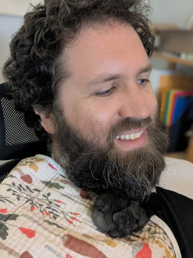 Alex Saines, Chief Financial Officer at Mycelium Creations, smiling in a portrait featuring a patterned woodland fabric. There is a black baby chick tucked under his chin.