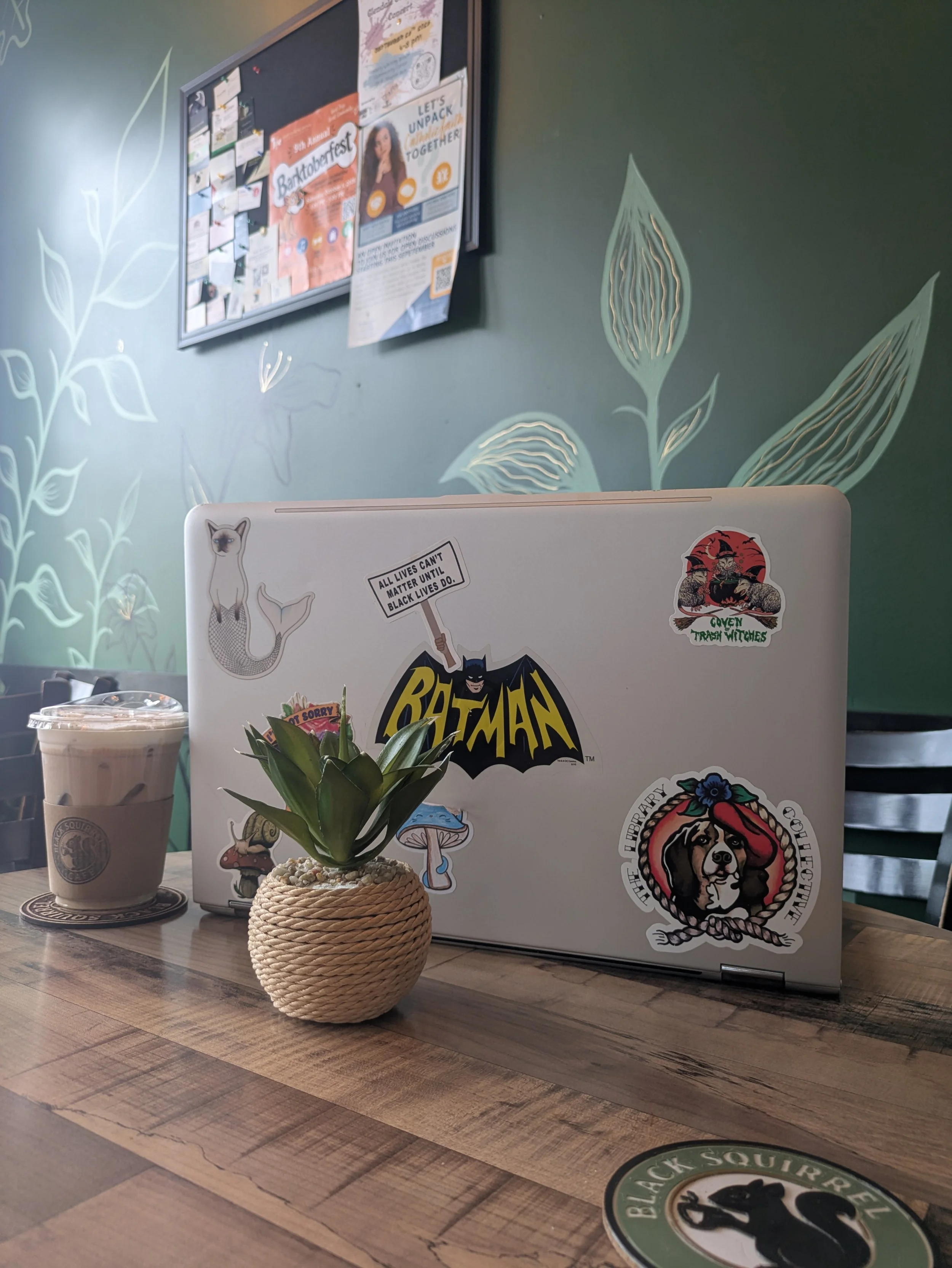 Silver laptop decorated with a vintage Batman logo, mushroom, and message stickers on a cafe table at Black Squirrel Cafe, in Glendale, Ohio, alongside an iced latte and a potted succulent with a green painted floral wall in the background.