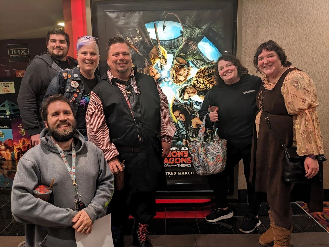 Cassie and her long-running tabletop roleplaying group smiling together in fantasy costumes in front of a Dungeons and Dragons movie poster.