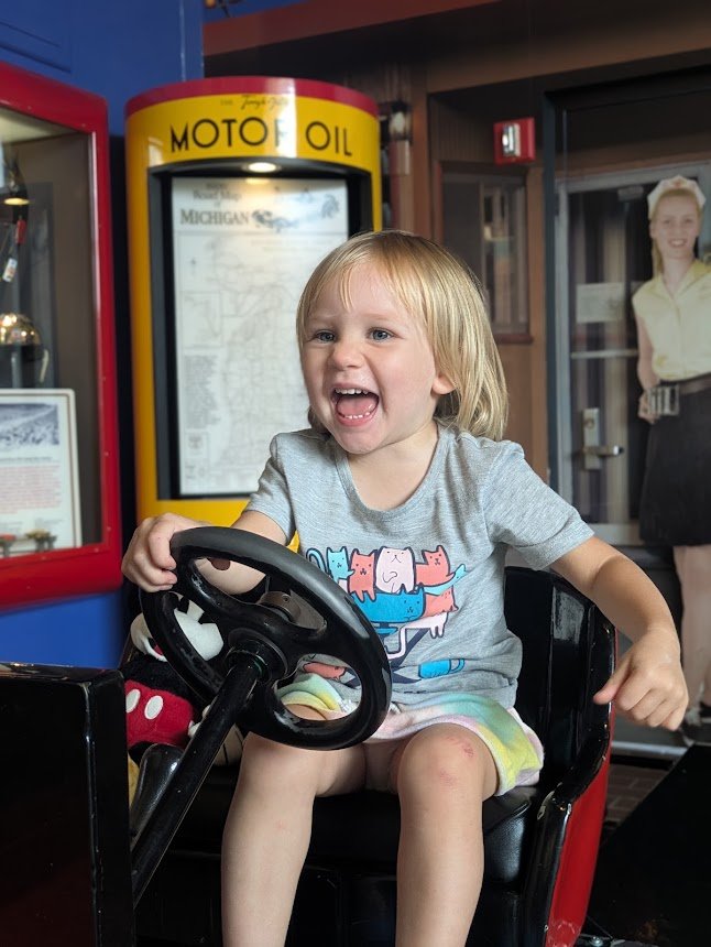 Cheerful young child holding a large steering wheel in an amusement display and laughing with open mouth.