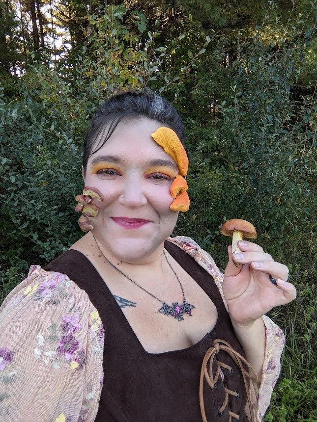 Cassie Saines, founder of Mycelium Creations, in a cottagecore dryad cosplay with mushroom face makeup holding a wild mushroom.