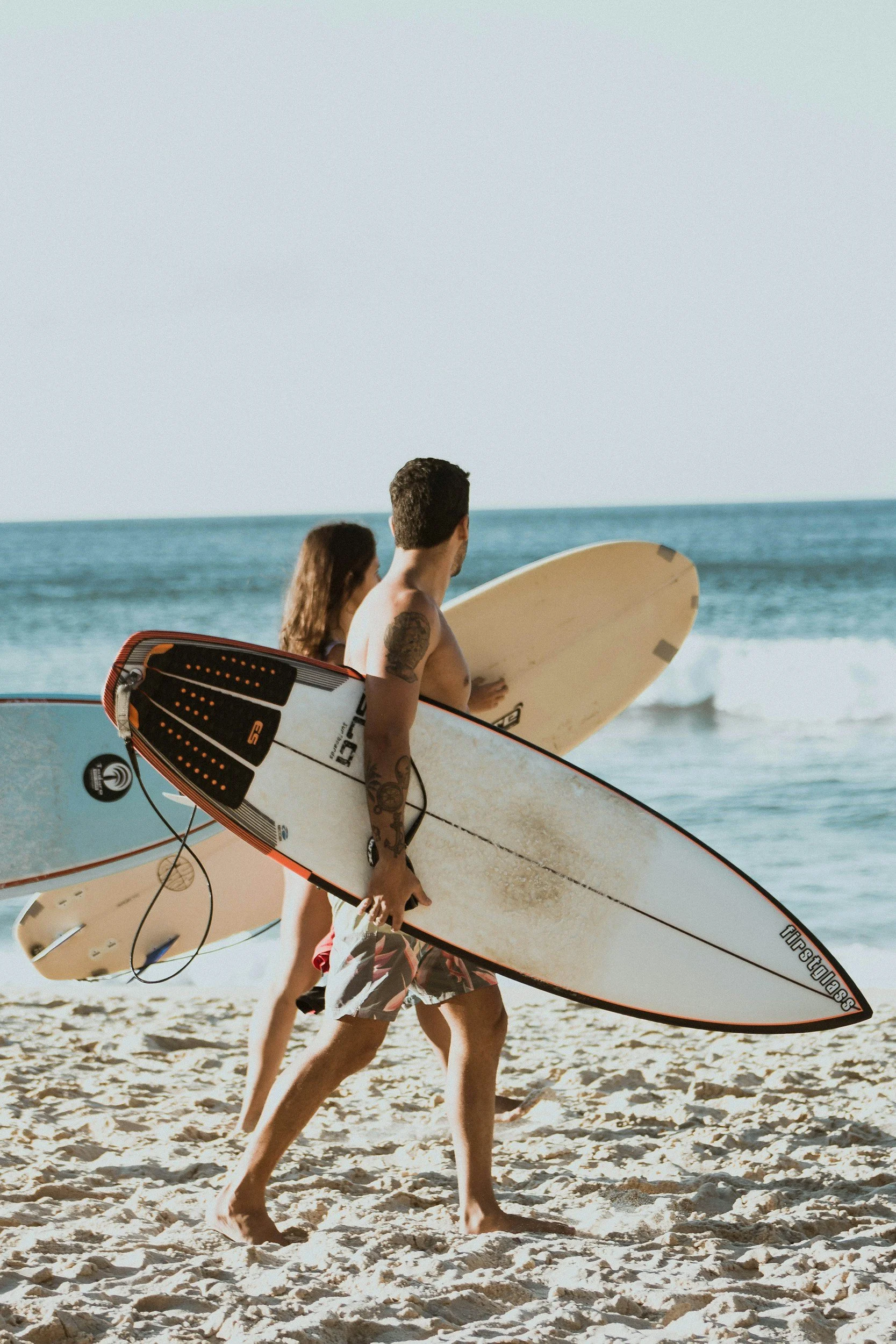 Two young adults, a male and a female, walking on a sandy beach carrying surfboards. The man has tattoos on his left arm and is shirtless, while the woman has long hair and is wearing a bikini top. They are heading towards the ocean with waves in the background.