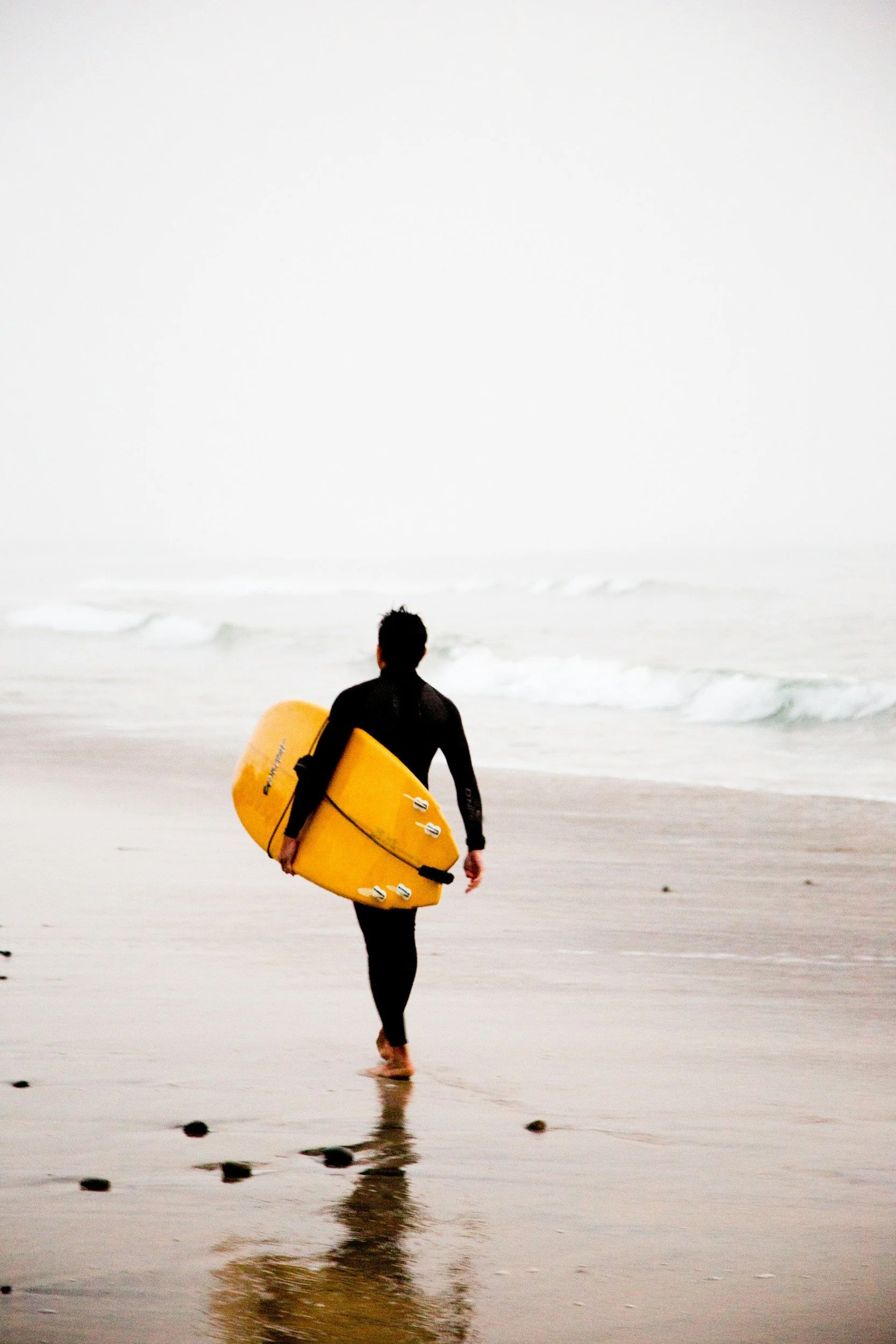 A person holding a yellow surfboard walks along the wet sand at the beach, facing the ocean, with overcast sky.