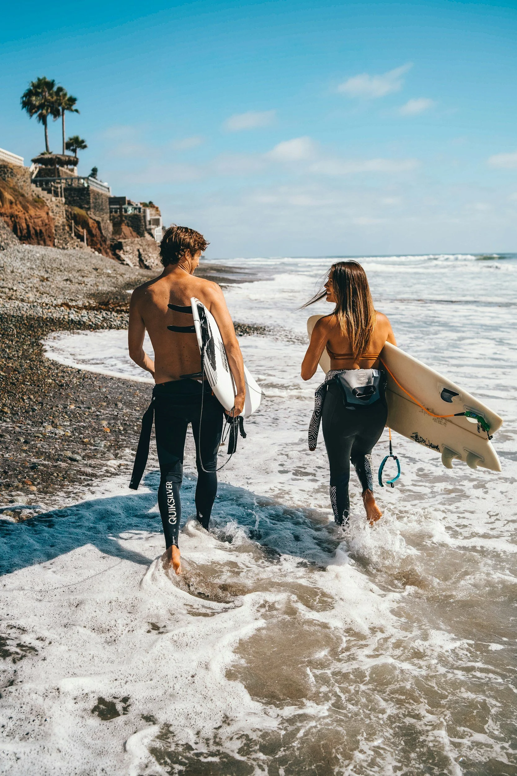 A man and a woman walking into the ocean waves on a beach, carrying surfboards, with houses and palm trees in the background under a blue sky.